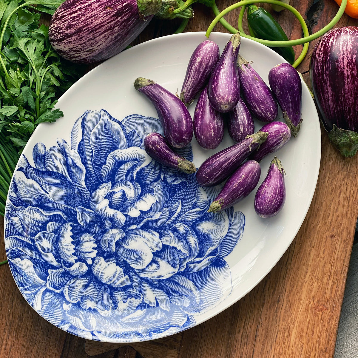 A decorative Peony Medium Coupe Oval Platter plate holding small purple eggplants, surrounded by various vegetables on a wooden surface.