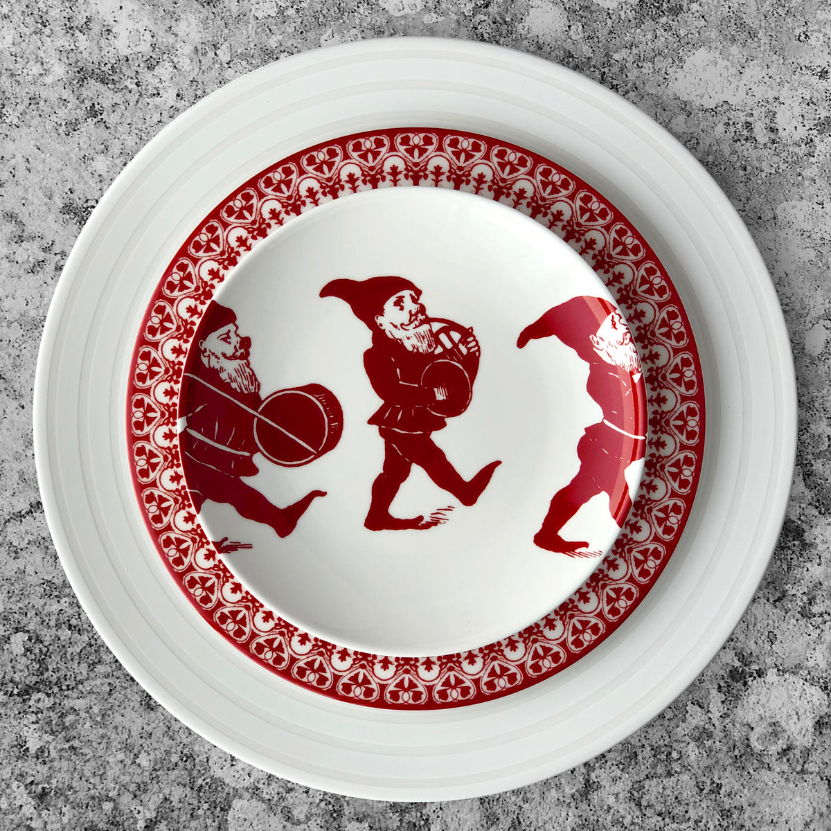 A white plate with a red decorative rim featuring three gnome-like figures playing musical instruments. The **Elves Small Plate** by **Caskata Artisanal Home**, part of an heirloom-quality dinnerware set, is placed on a larger white plate on a textured gray surface.