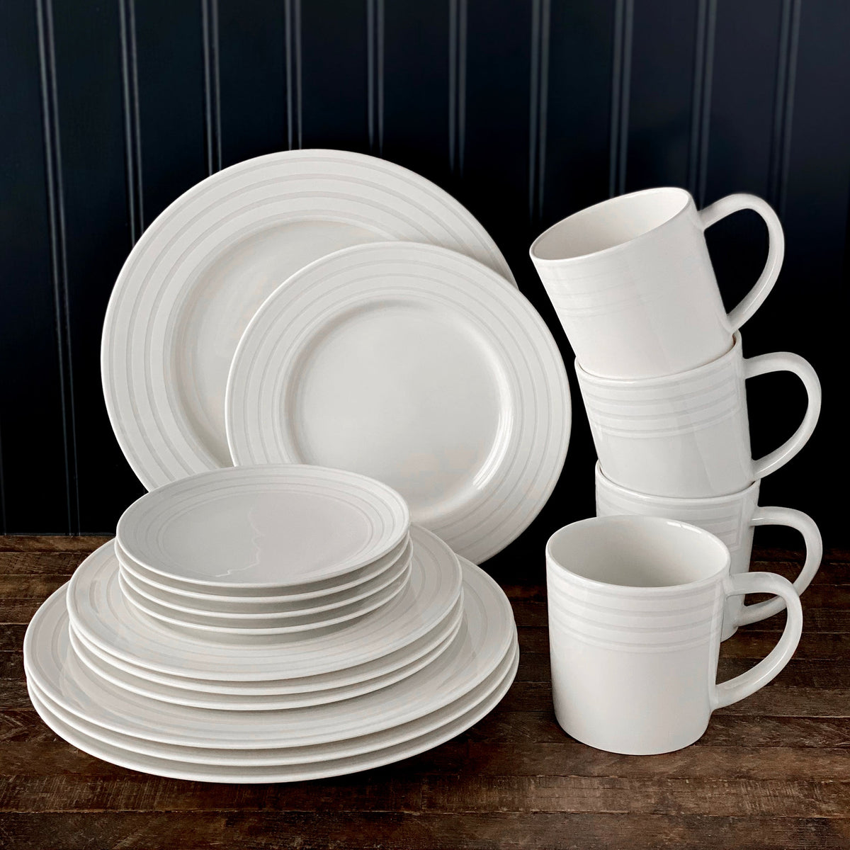 A neatly stacked set of white dishware, including Caskata Artisanal Home Cambridge Stripe Rimmed Salad Plates, bowls, and mugs, arranged against a dark-paneled background on a wooden surface.