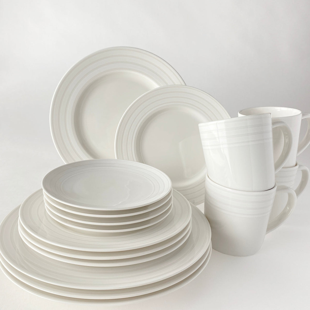A set of white ceramic dinnerware, including Caskata Artisanal Home Cambridge Stripe Small Plates, bowls, and mugs with a subtle Cambridge Stripe pattern, arranged neatly against a plain background.