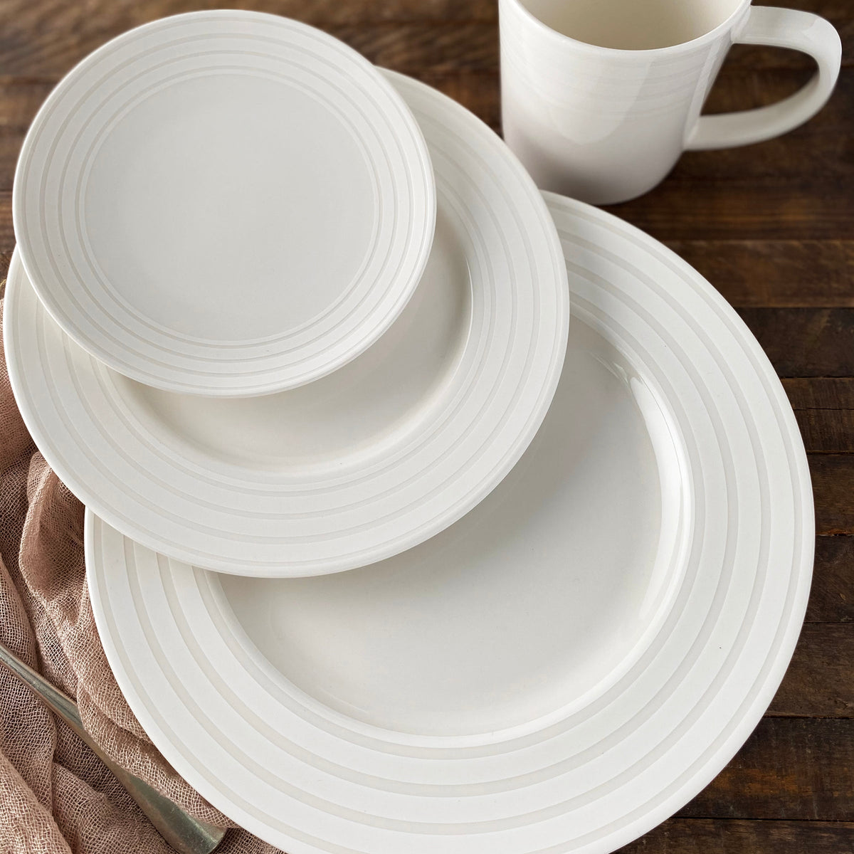 A white ceramic dinner set consisting of a large plate, a medium plate, Cambridge Stripe Small Plates by Caskata Artisanal Home, and a mug on a wooden surface. A pink cloth and a fork are partially visible, creating an inviting table setting.