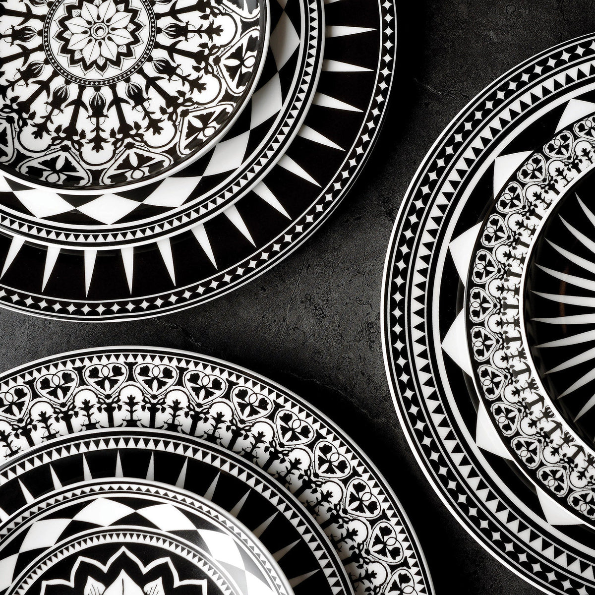 A collection of Casablanca Rimmed Dinner Plates by Caskata Artisanal Home featuring black and white ceramic plates with intricate geometric and floral patterns, all meticulously hand-decorated, arranged on a dark surface.