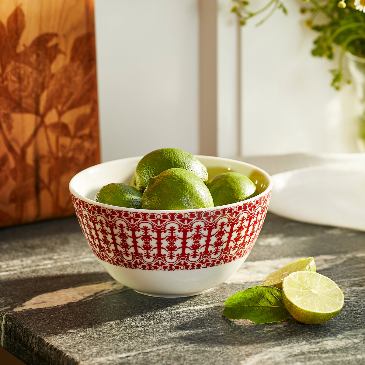 A **Casablanca Crimson Cereal Bowl by Caskata**, filled with green limes sits on a kitchen countertop near a halved lime and a basil sprig. Its high-fired porcelain finish adds an elegant touch to the scene.