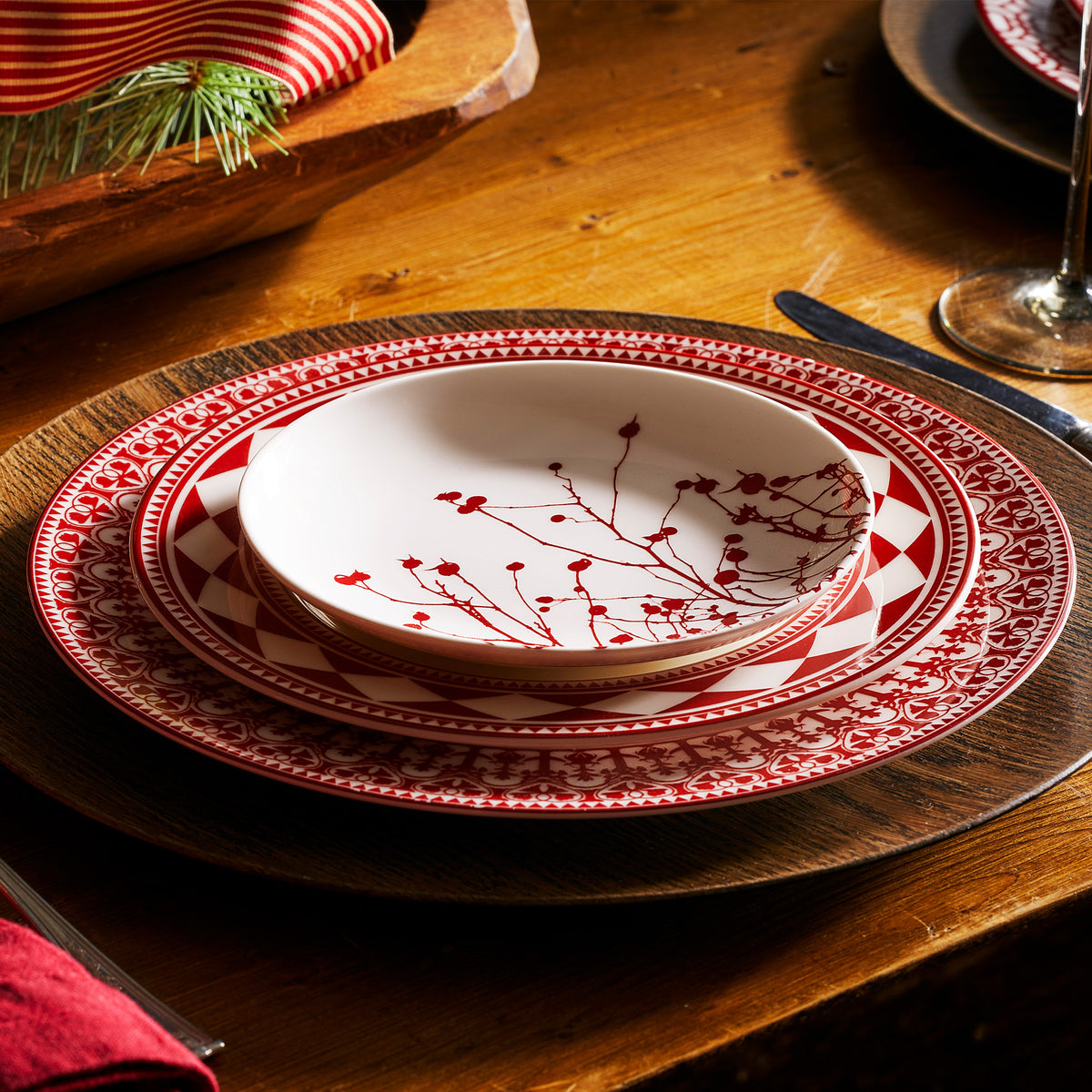 A wooden table set with heirloom-quality dinnerware showcasing a decorative plate arrangement featuring red and white patterns and branch designs, accompanied by a knife and a fork. The Caskata Artisanal Home Winterberries Small Plates add an elegant touch, reminiscent of Winter berries.