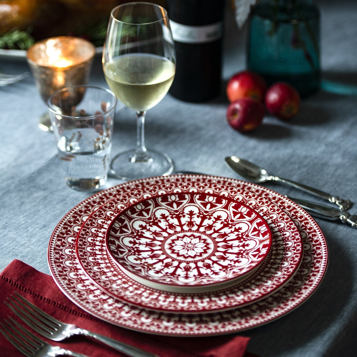 A dining table set with heirloom-quality Caskata Artisanal Home Casablanca Crimson Small Plates, featuring red and white patterned plates, a glass of white wine, a water glass, cutlery, a red napkin, and a candle in the background.