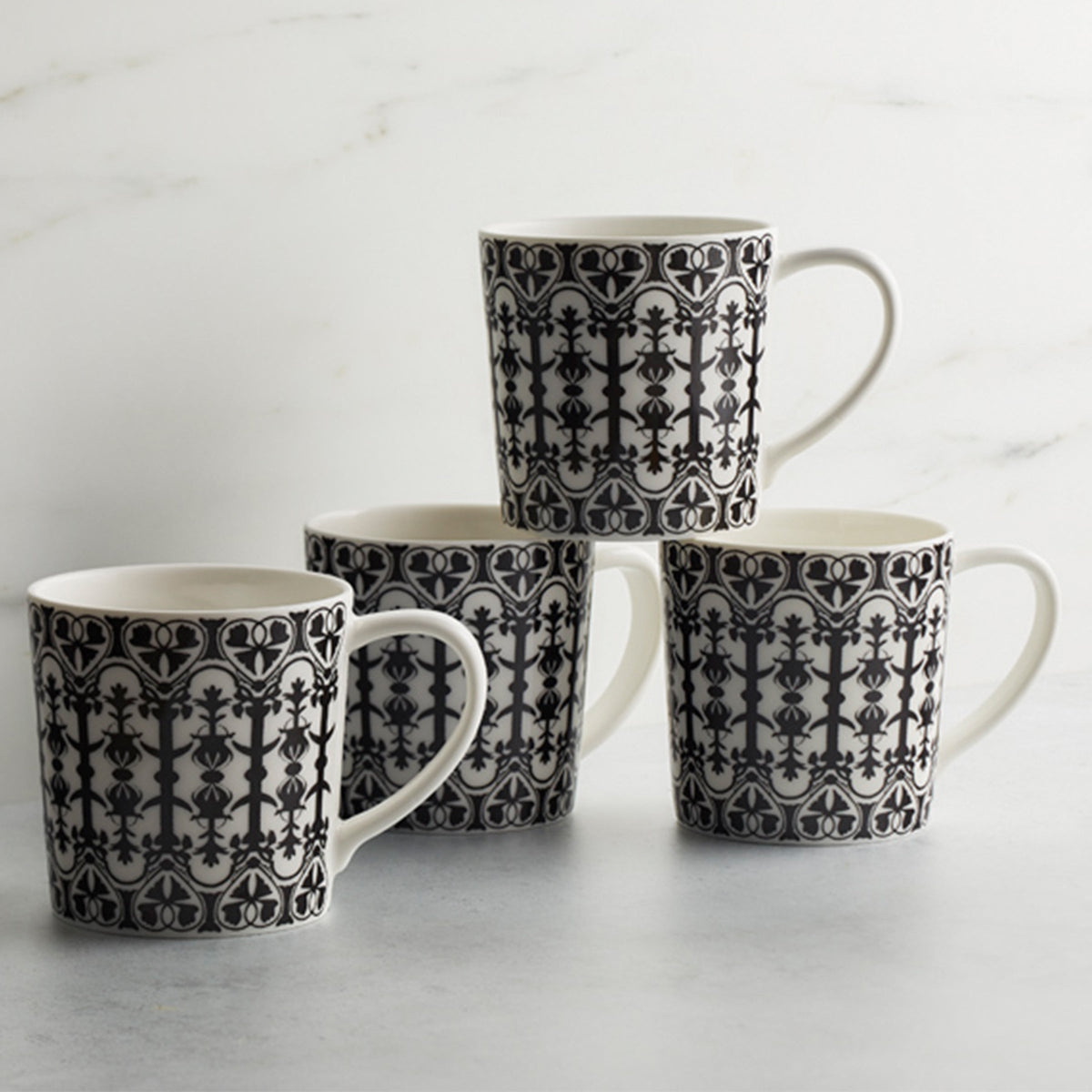 Four Casablanca Mugs by Caskata Artisanal Home, with black intricate geometric and floral patterns, arranged in a square formation on a light gray surface against a white marble backdrop, echo the exquisite charm of Casablanca dinnerware.