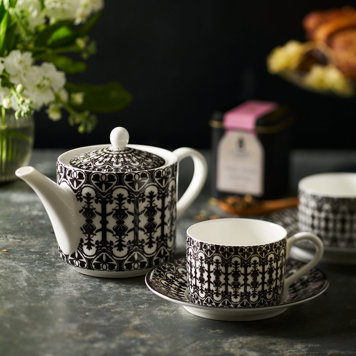 A black and white patterned teapot with matching Casablanca Cups &amp; Saucers, Set of 2 by Caskata is placed on a table. Nearby are flowers in a vase, a tin of tea, and a plate of baked goods, creating an inviting scene with elegant dinnerware.