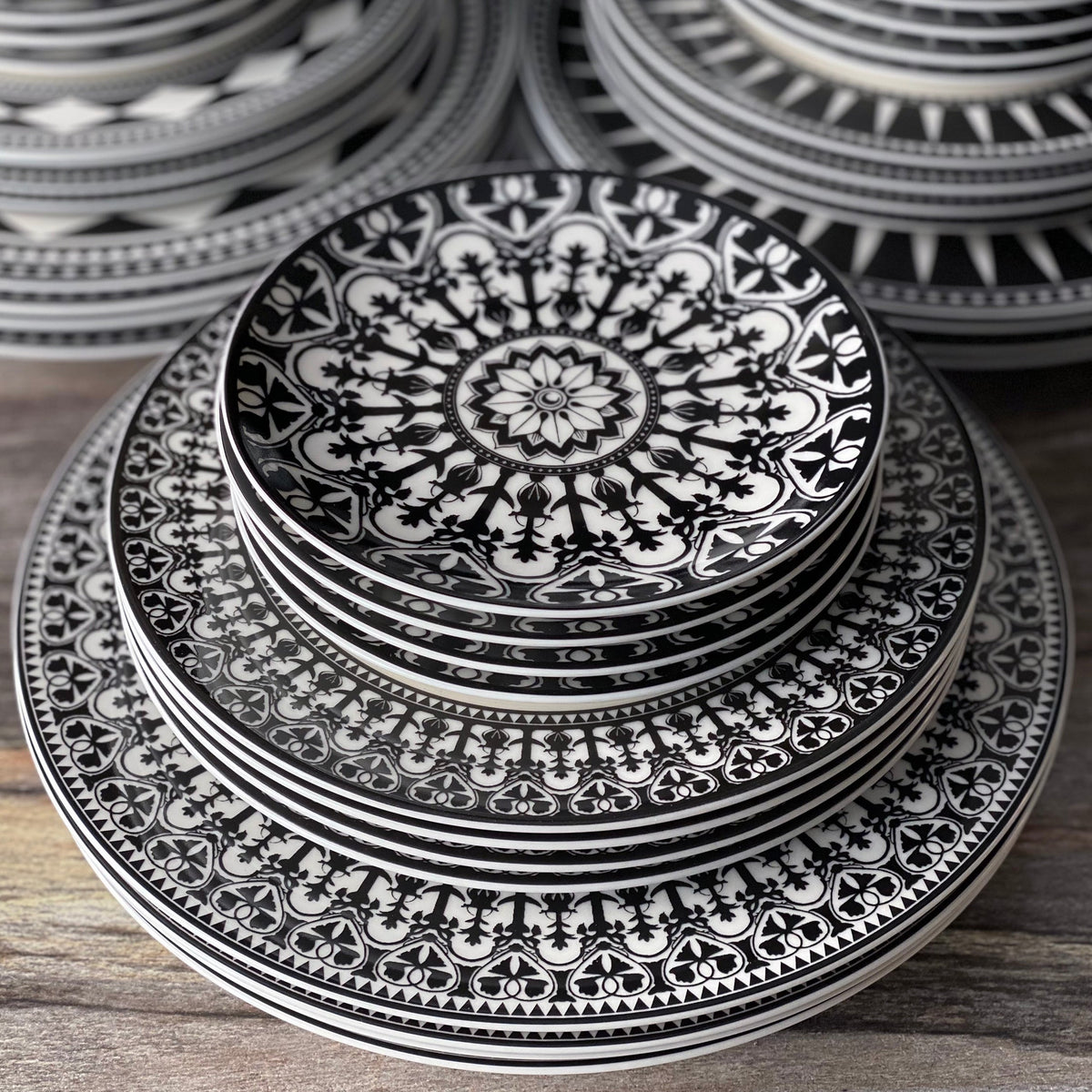 A stack of Casablanca Rimmed Dinner Plates by Caskata Artisanal Home with intricate black and white geometric and floral patterns, hand-decorated details reminiscent of Casablanca dinnerware, arranged in size order on a wooden surface.
