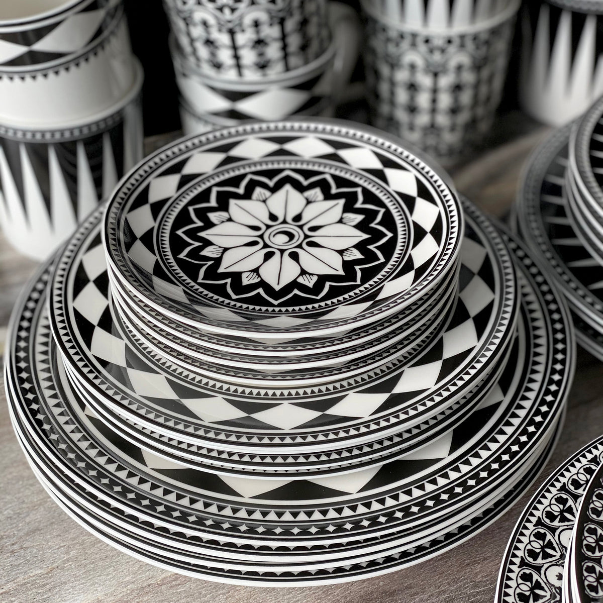 A stack of black and white patterned Fez Rimmed Dinner Plates by Caskata Artisanal Home of varying sizes, with matching bowls and cups in the background, showcases sophisticated dinnerware with a touch of Moroccan patterns.