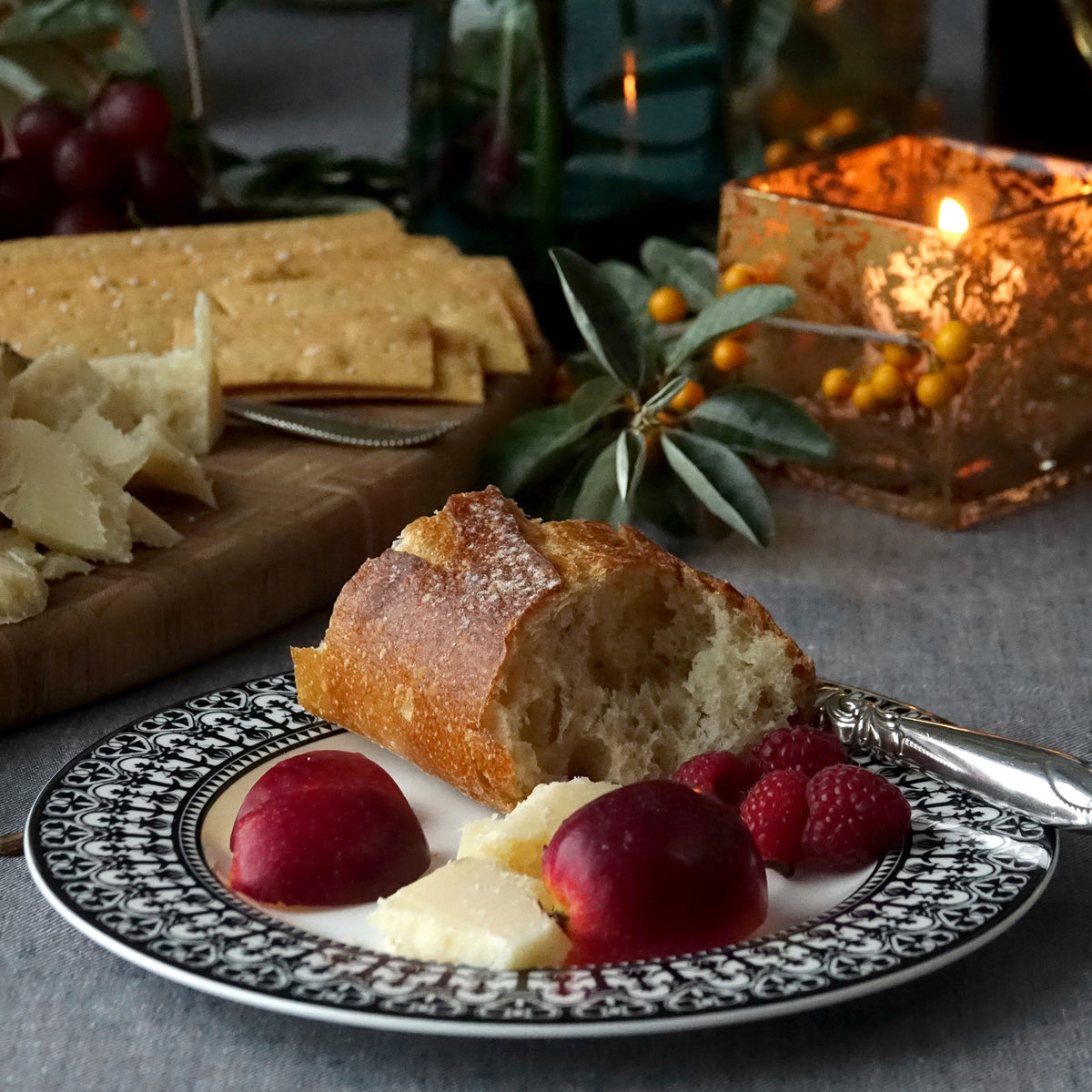 A plate with a chunk of bread, cheese, plums, and raspberries, set on an exquisite Casablanca Rimmed Salad Plate by Caskata Artisanal Home adorned with hand-decorated details. In the background, a candle, grapes, and crackers complete the inviting scene.