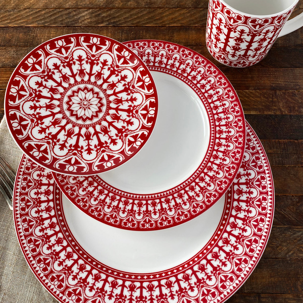 A table setting with heirloom-quality white and red patterned Casablanca dinnerware, including Caskata Artisanal Home Casablanca Crimson Small Plates, salad plate, bowl, and cup arranged neatly on a wooden surface.