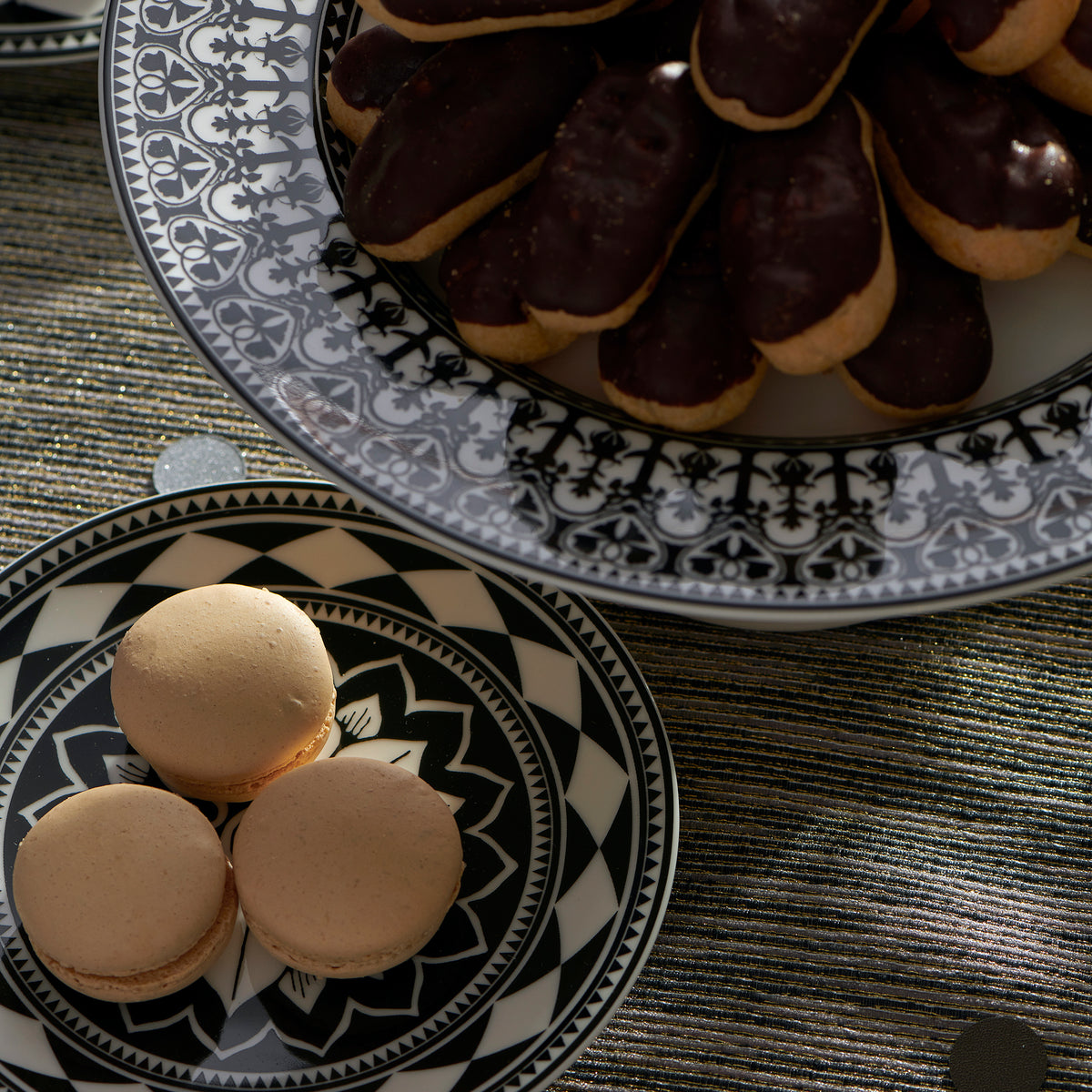 A plate of chocolate-covered eclairs is placed on a larger ornate plate, adorned with Moroccan patterns, while three beige macarons are positioned on a smaller decorative Caskata Artisanal Home Fez Small Plates nearby.