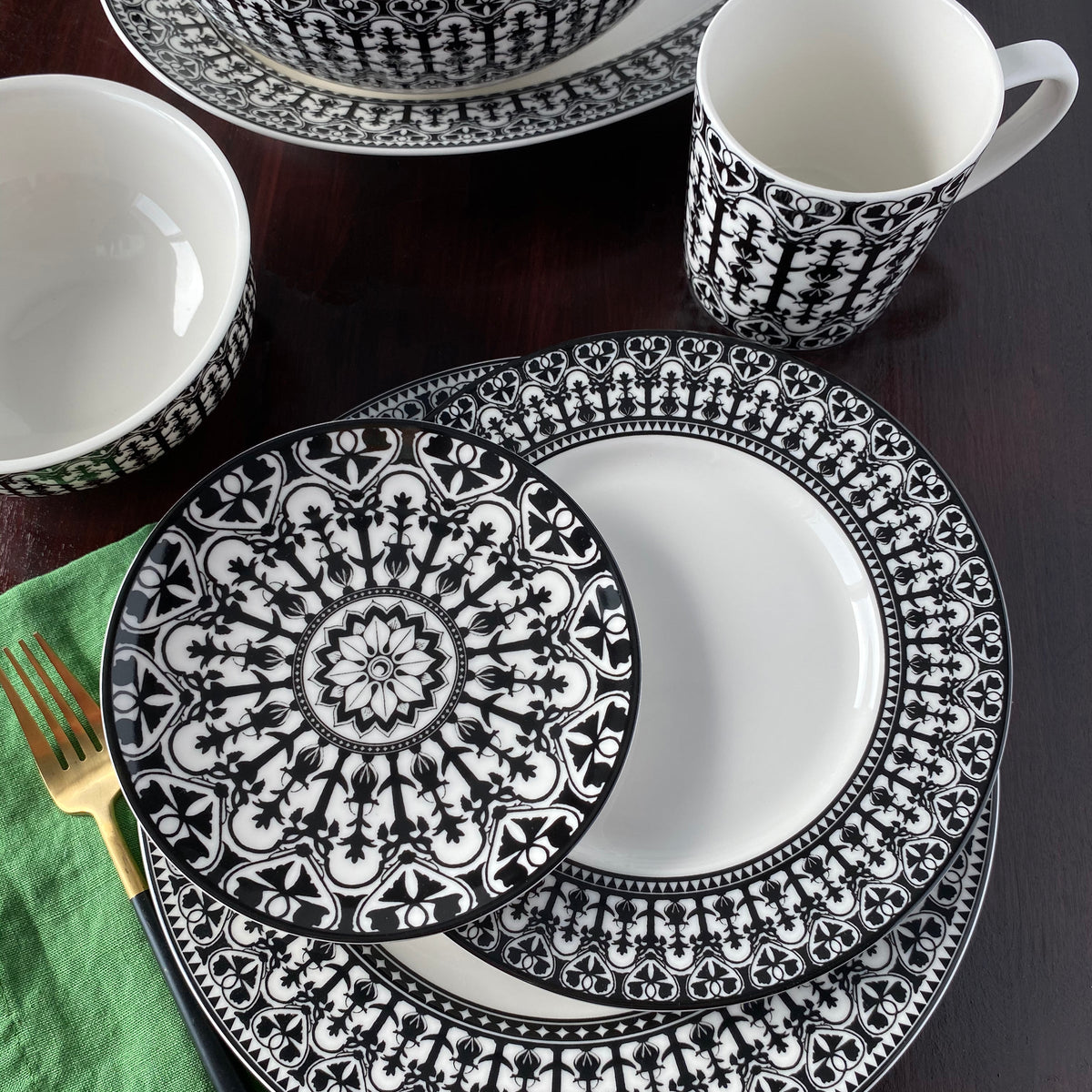 A Casablanca Rimmed Dinner Plate by Caskata Artisanal Home featuring a black and white patterned design is meticulously arranged on a dark wood table with a green napkin and gold fork. Made from high-fired porcelain, the plate is part of a set that includes bowls and a mug.