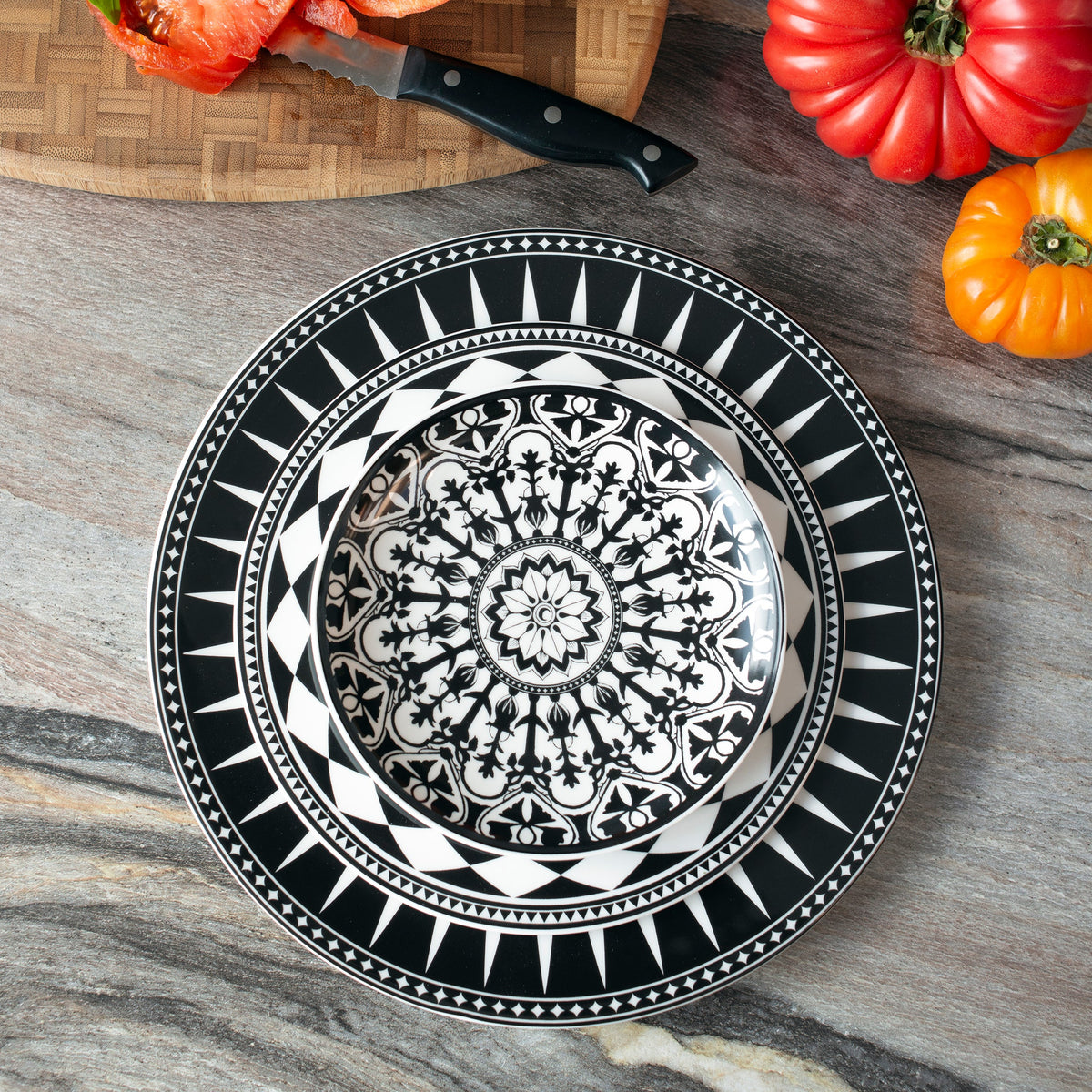 A Caskata Artisanal Home Marrakech Rimmed Dinner Plate with intricate patterns, reminiscent of Marrakech dinnerware, is displayed on a countertop surrounded by colorful vegetables and a cutting board with a knife.