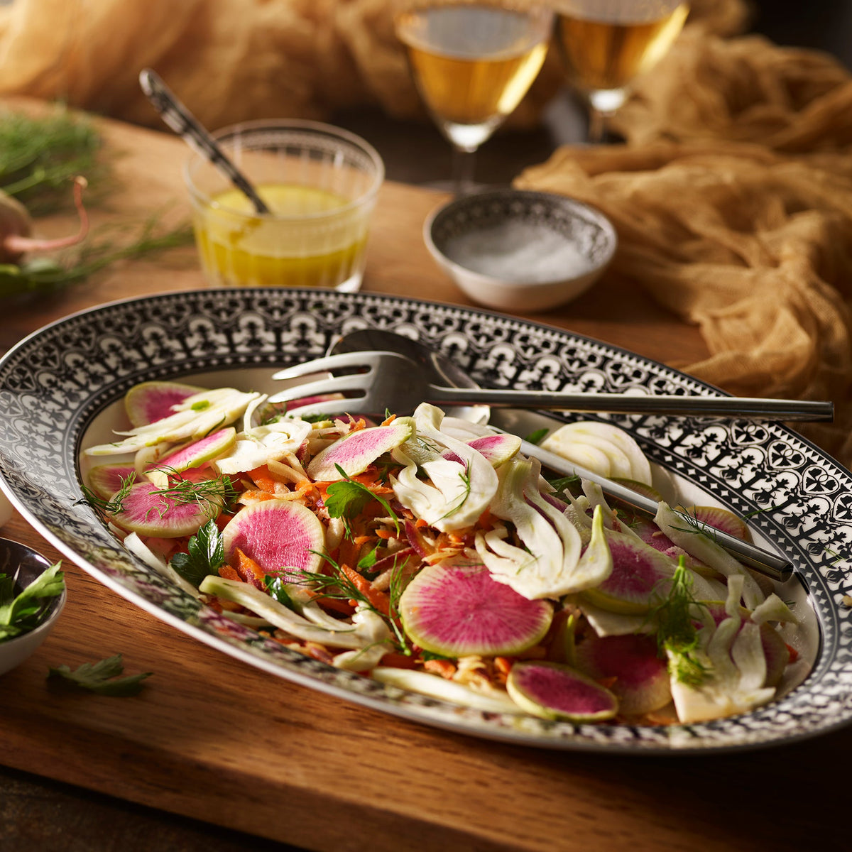 A Caskata Artisanal Home Casablanca Oval Rimmed Platter adorned with ornate scrollwork holds a fresh salad with sliced radishes, fennel, and herbs. A fork rests on the dish. Bowls of dressing and salt, along with two glasses of white wine, are in the background.