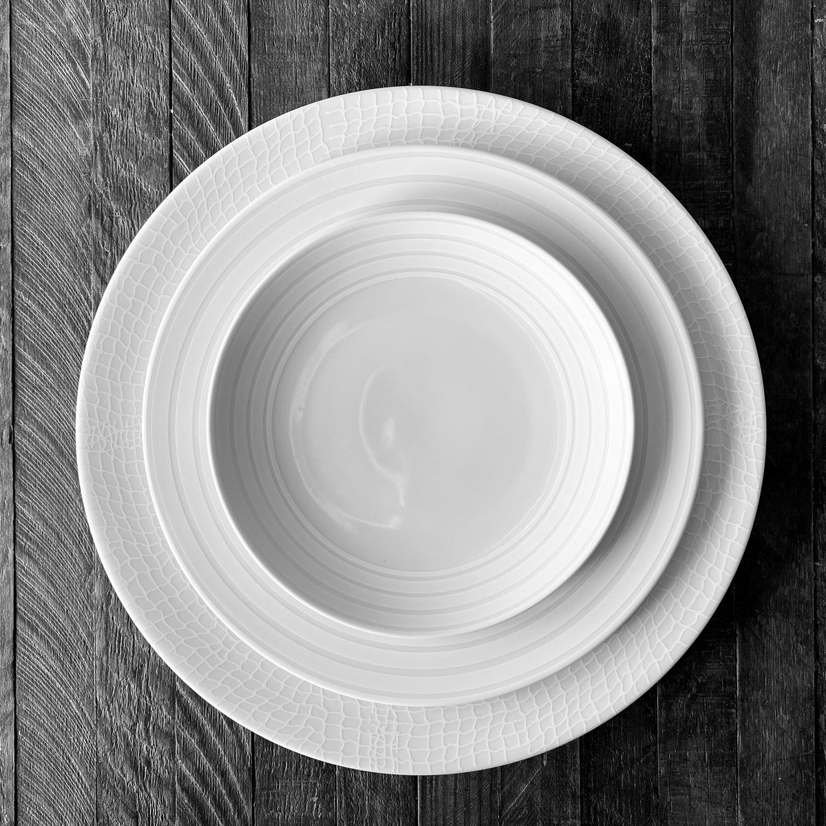 A high-fired porcelain bowl neatly placed inside a Cambridge Stripe Rimmed Salad Plate by Caskata Artisanal Home, which in turn rests on a larger white dinner plate, all elegantly arranged on a dark wooden surface.
