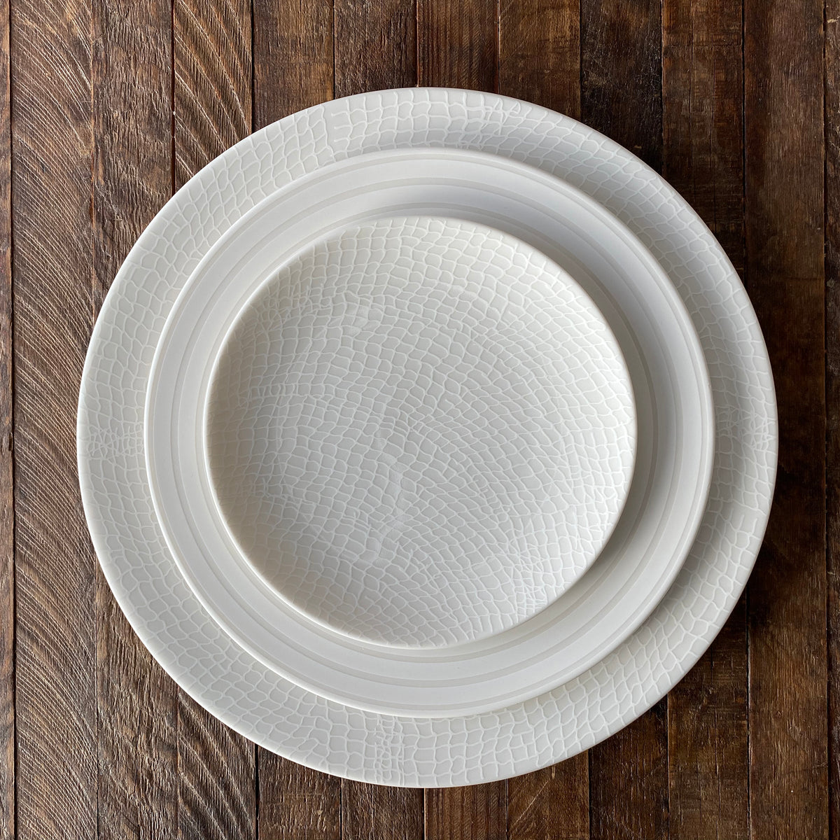 Two stacked Catch Small Plates by Caskata Artisanal Home made of high-fired porcelain with a textured, white design are placed on a wooden surface.