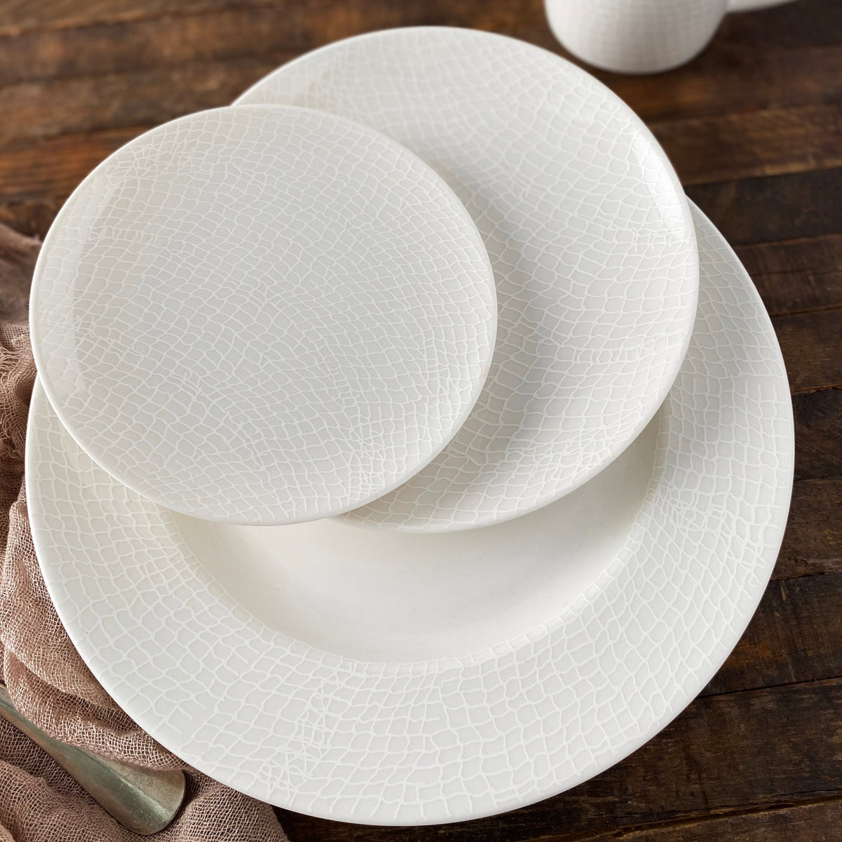 A set of three Catch Small Plates by Caskata Artisanal Home, with a textured pattern, arranged on a wooden table alongside a light brown cloth, evokes the intricate design of a fisherman&#39;s net.