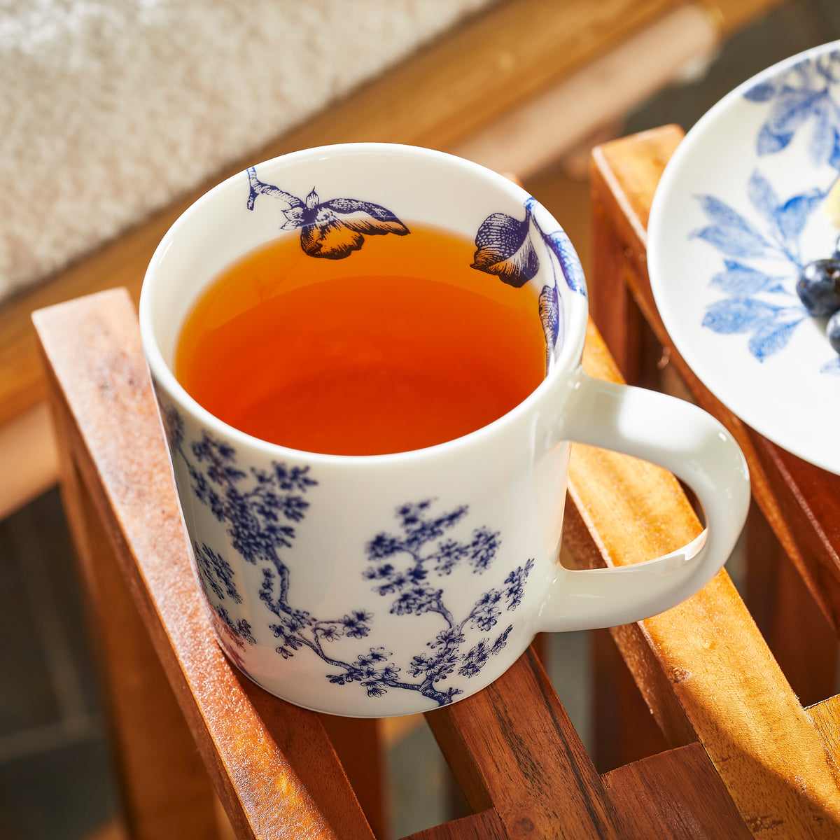 A Chinoiserie Toile Mug by Caskata Artisanal Home filled with tea is placed on a wooden surface next to a white plate with a matching Chinoiserie Toile pattern, embodying timeless elegance.