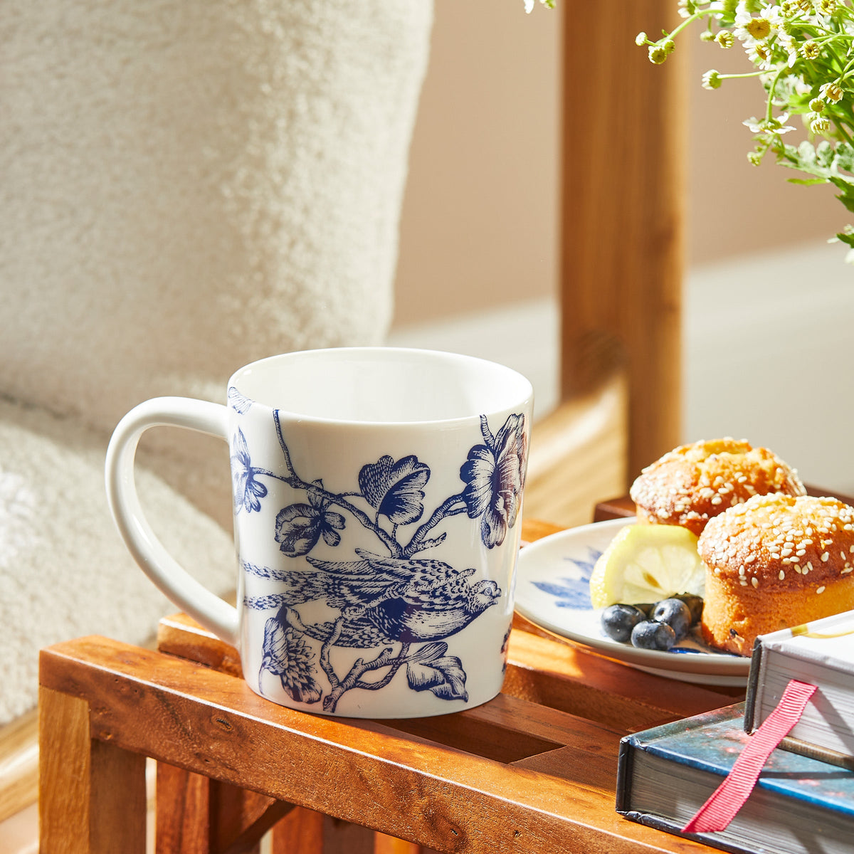 A **Chinoiserie Toile Mug** by **Caskata Artisanal Home**, embodying timeless elegance, sits on a wooden tray beside a plate of muffins, blueberries, and a slice of lemon. Nearby, there are several stacked books and a vase with green foliage.