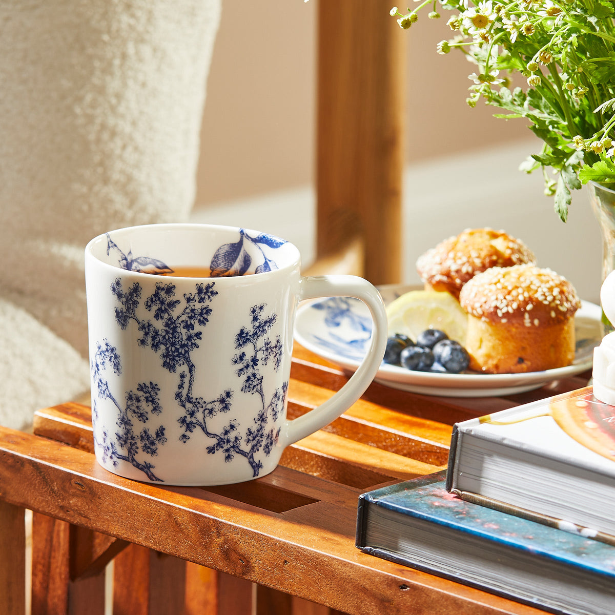 A **Chinoiserie Toile Mug** by **Caskata Artisanal Home**, reminiscent of Chinoiserie Toile, sits on a wooden surface next to books, a plant, and a plate with muffins, blueberries, and lemon slices.