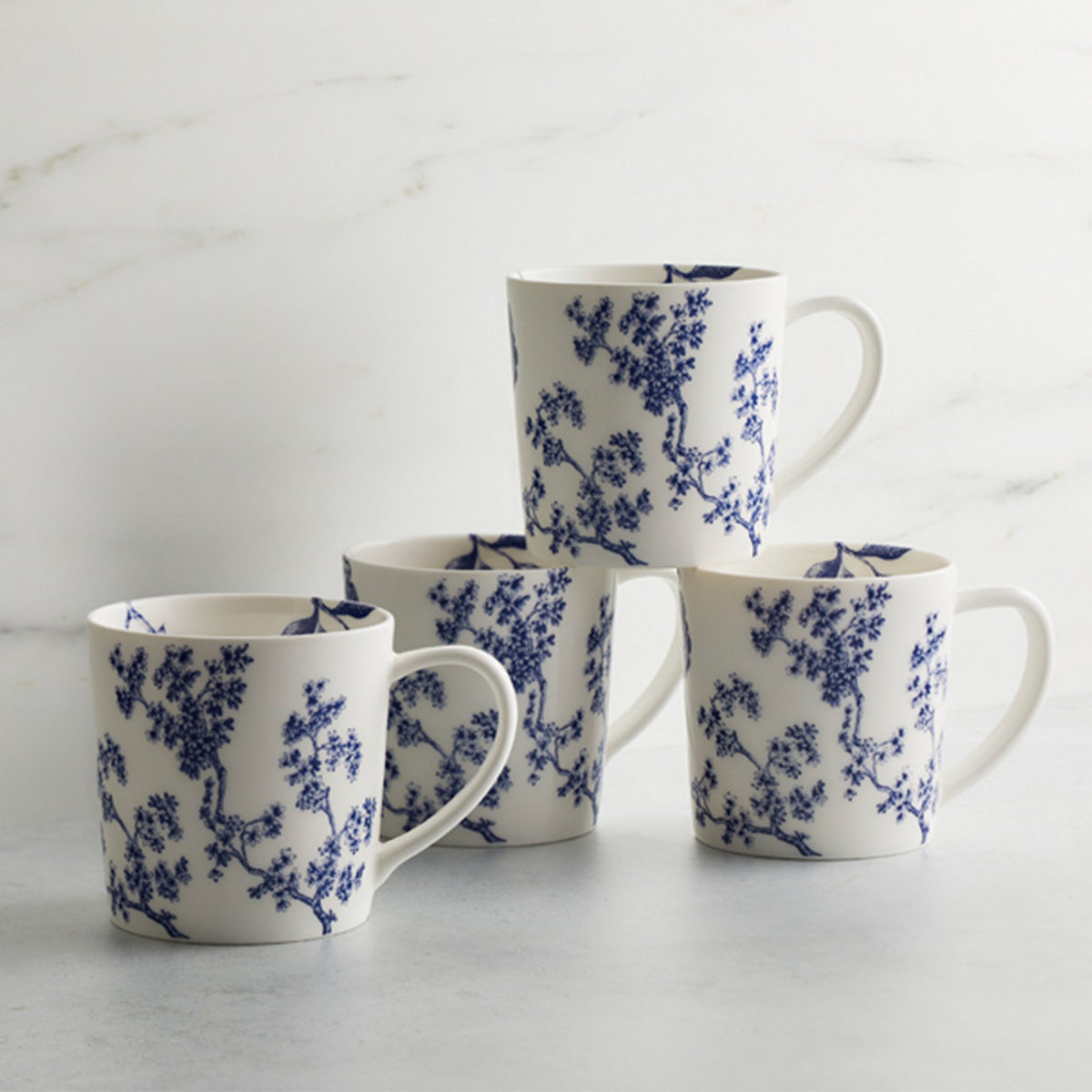 Four Chinoiserie Toile Mugs by Caskata Artisanal Home, with blue floral patterns, reminiscent of Chinoiserie Toile, are stacked and arranged on a white surface against a light, marbled background.