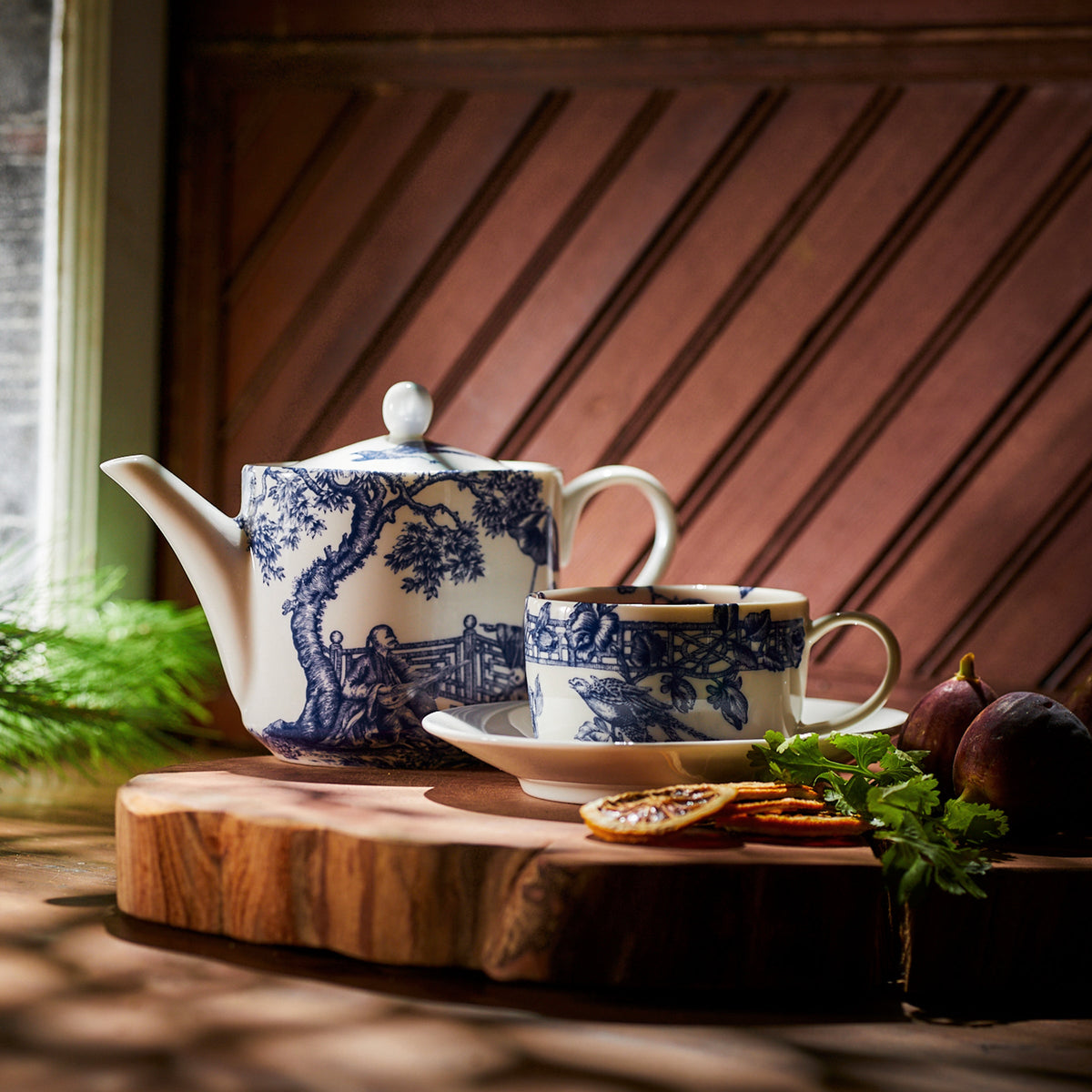 A Chinoiserie Toile teapot and Caskata Artisanal Home Chinoiserie Toile Cups &amp; Saucers, Set of 2 with blue-and-white patterns sit on a wooden board with figs and sliced dried fruit, set against a warmly lit wooden backdrop.