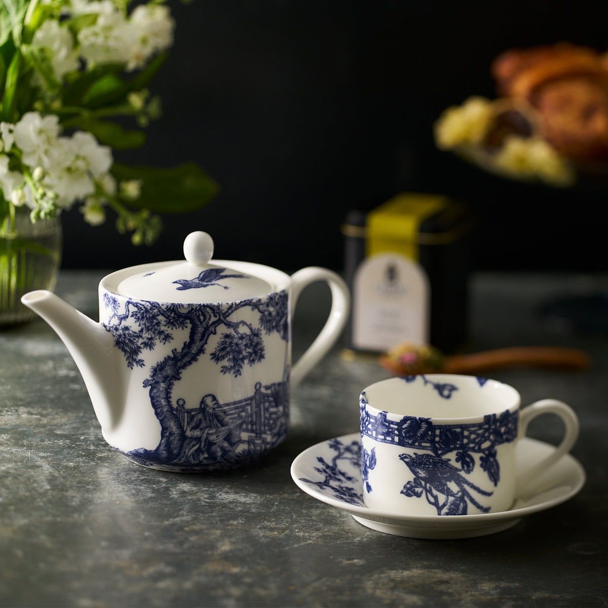 A stunning Chinoiserie Toile blue and white porcelain teapot and **Caskata Artisanal Home Chinoiserie Toile Cups &amp; Saucers, Set of 2** adorned with tree and bird designs are on a stone countertop. They are beside a bouquet of flowers, a tin of tea, and a plate of pastries in the background.