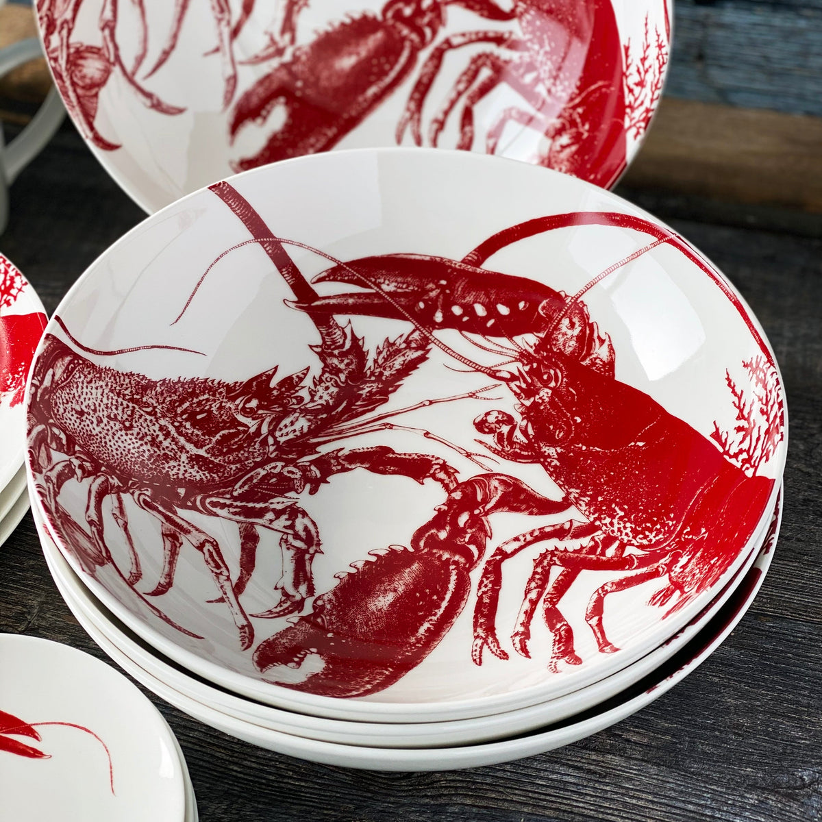 A stack of Lobster Wide Serving Bowls by Caskata Artisanal Home with red lobster illustrations on them, placed on a dark surface, perfect for adding a touch of seaside style to your tableware.