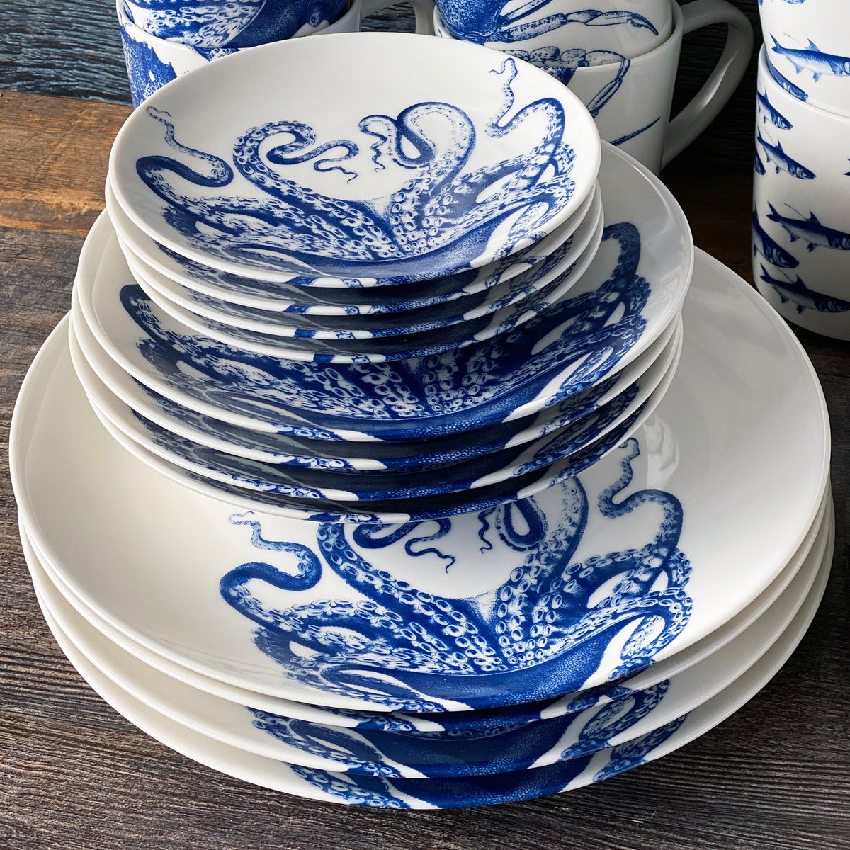 Stacked creamy white Lucy Coupe Dinner Plates by Caskata Artisanal Home with blue octopus designs, arranged on a wooden surface, with matching mugs in the background.