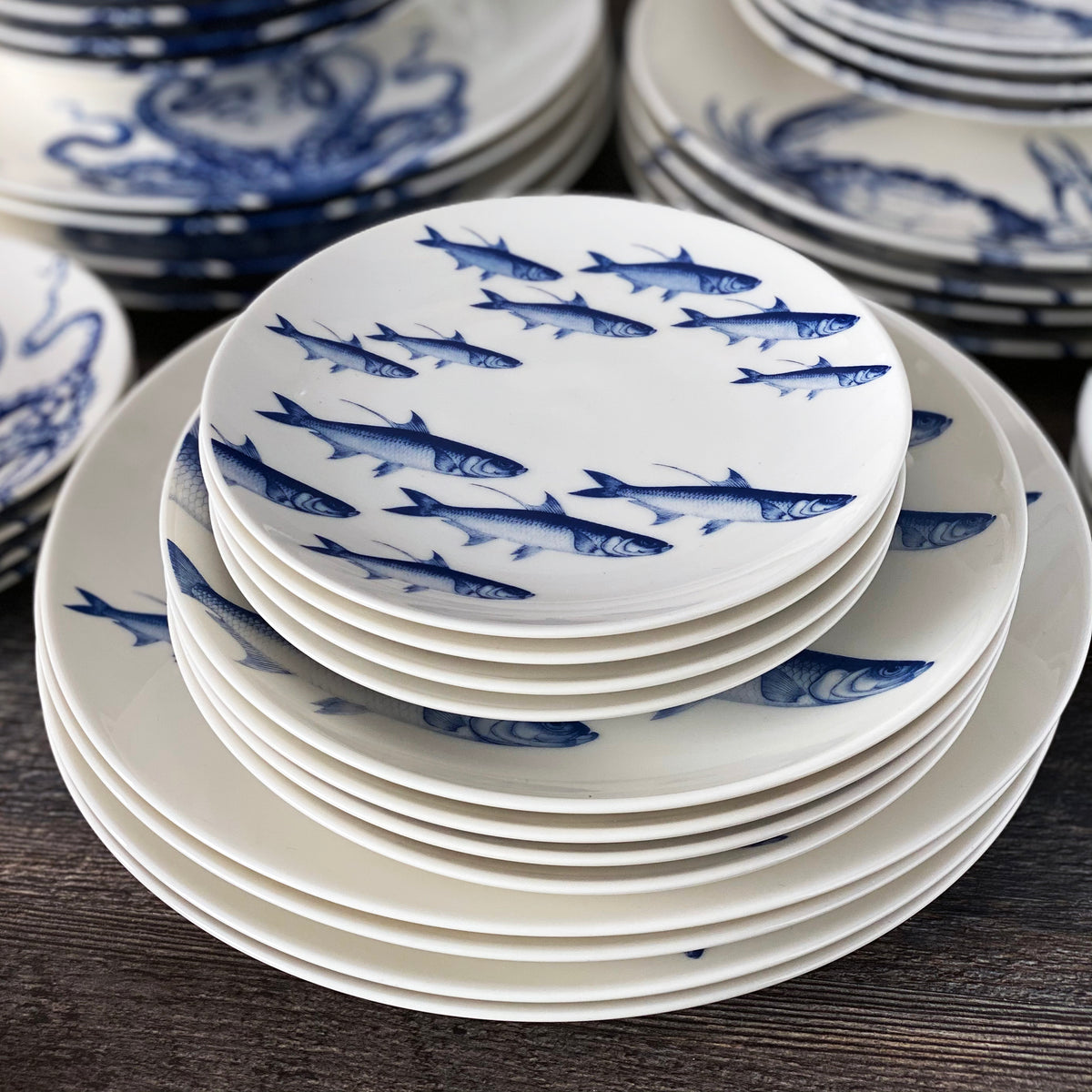 Stack of white ceramic plates decorated with blue fish patterns arranged on a dark wooden surface. These Caskata Artisanal Home School of Fish Small Plates, featuring a school of fish design, are also microwave safe for your convenience.