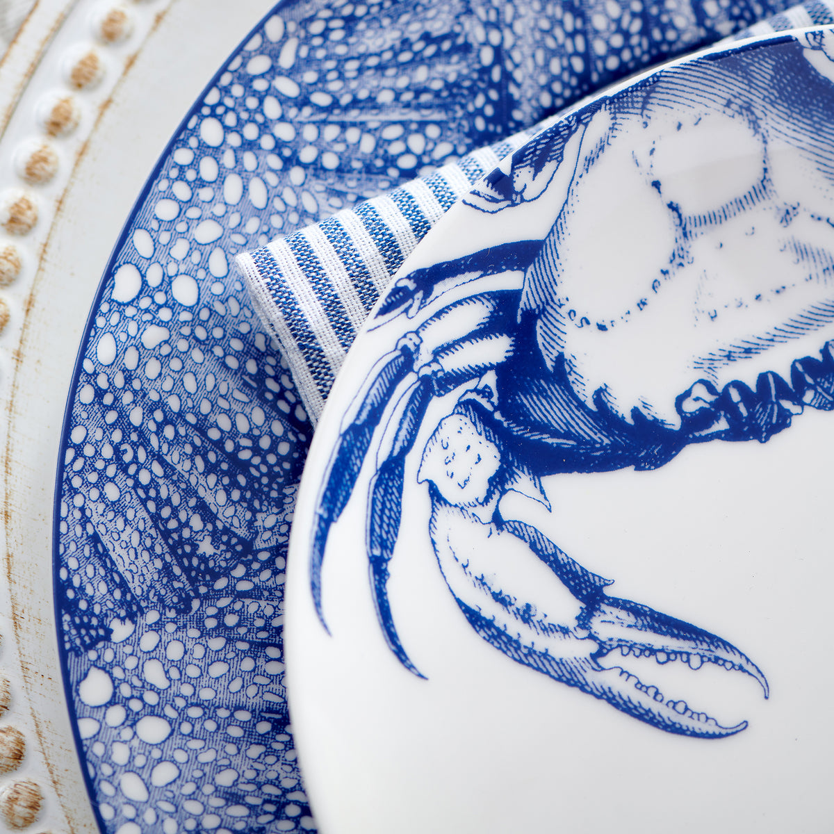 Close-up of a blue and white Crab Coupe Salad Plate by Caskata Artisanal Home featuring a crab pattern, styled with a striped napkin and placed on a decorative plate with a textured edge.