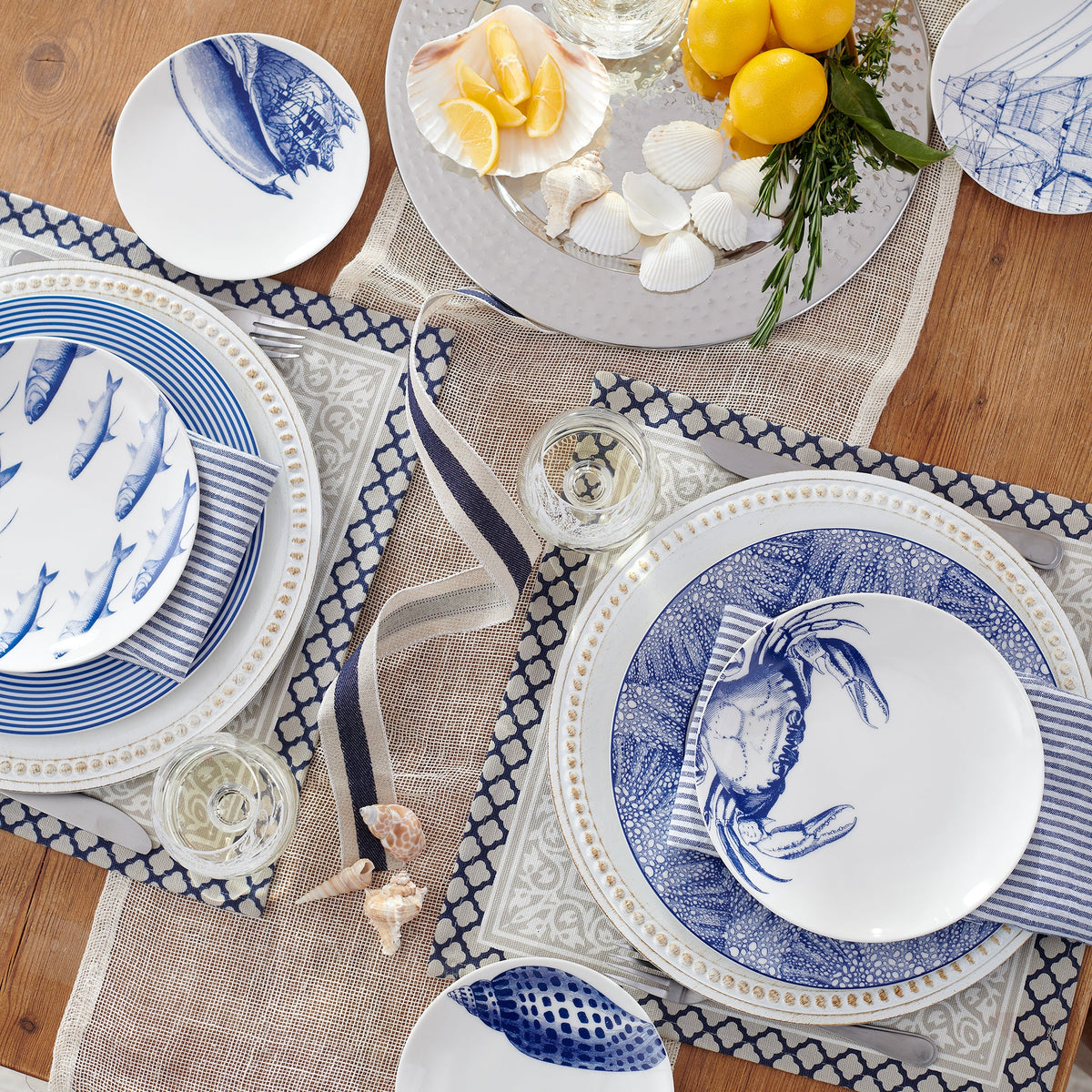 A wooden table set with coastal-themed Crab Coupe Salad Plates by Caskata Artisanal Home featuring premium porcelain and a charming crab pattern, surrounded by sea shells, lemons, and glasses, arranged on a mesh table runner.