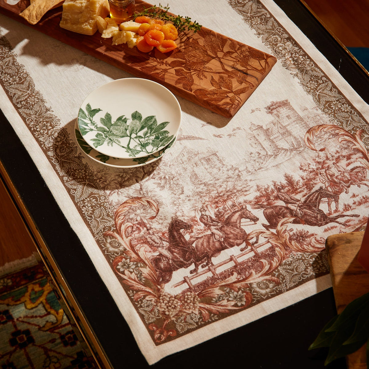A table is set with a vintage tablecloth featuring horse and carriage designs, a wooden cheese board with cheese and apricots, and Caskata&#39;s Arbor Green Small Plates adorned with a green leaf motif.