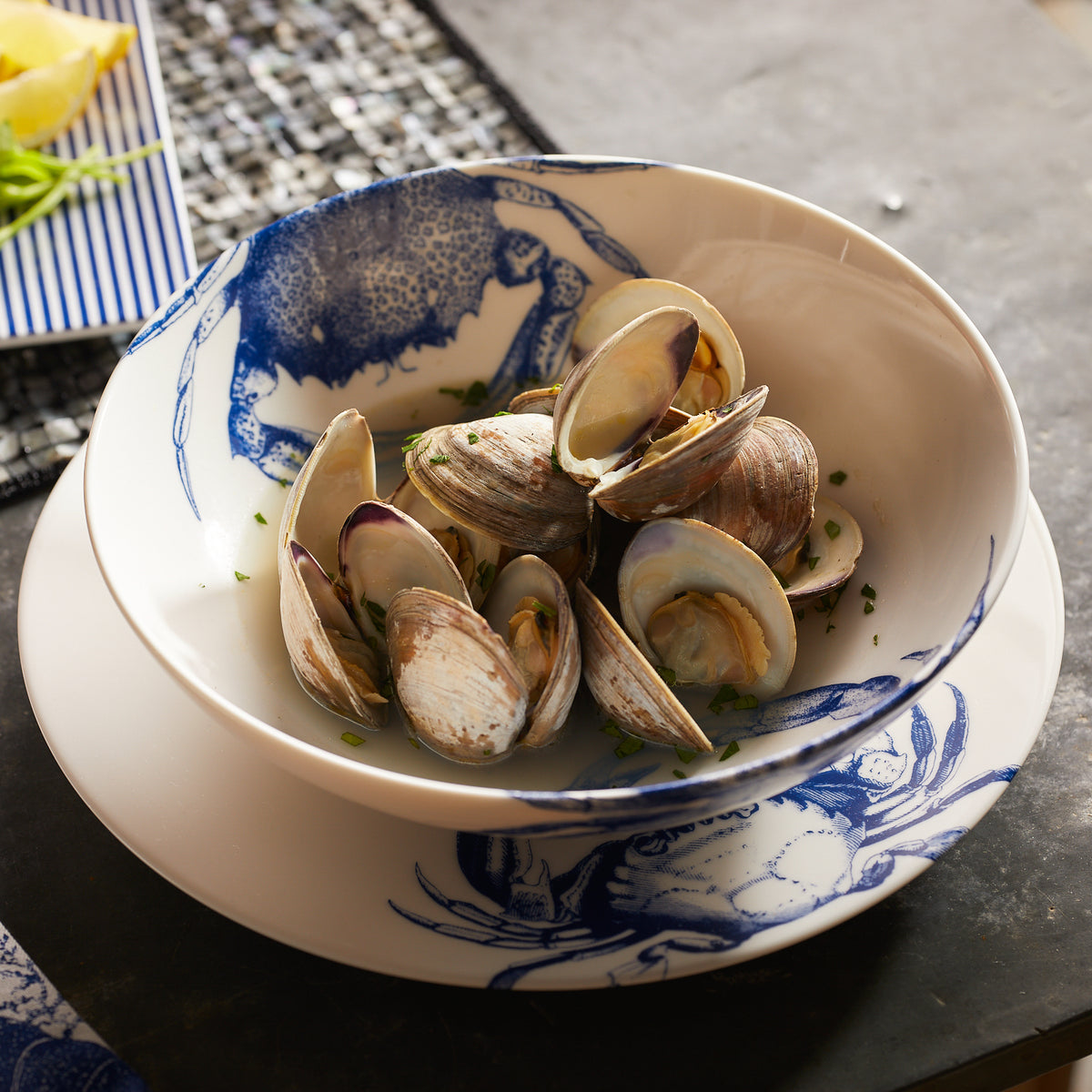 A bowl of cooked clams served in a white dish adorned with a charming crab pattern, placed on a matching plate with part of a lemon wedge visible in the background. This high-fired porcelain set, the Crab Entrée Bowl by Caskata Artisanal Home, is both dishwasher and microwave safe, ensuring convenience and durability.