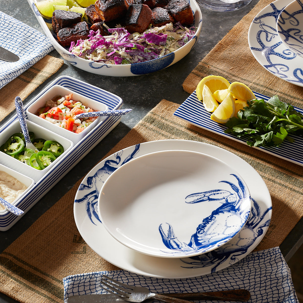 A table is set with Caskata Artisanal Home Crab Coupe Salad Plates made of premium porcelain, a dish of mixed salad and charred meat, a tray with condiments, sliced lemons, and herbs. A fork and knife rest on burlap placemats.