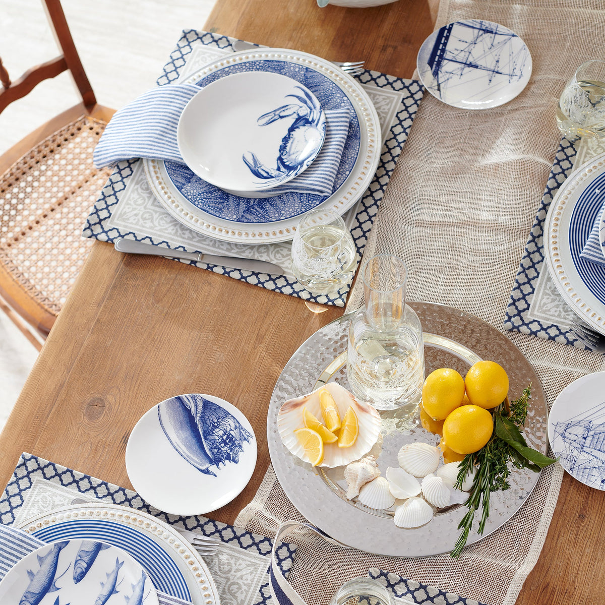 A wooden dining table set with heirloom-quality dinnerware, folded napkins, glassware, a silver platter with lemons, herbs, and Shells Small Plates from Caskata Artisanal Home&#39;s beach collection, and a mesh table runner.