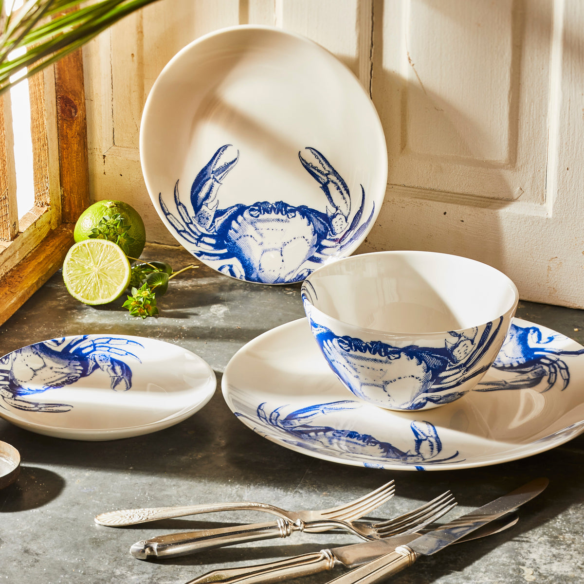 A premium porcelain set of white ceramic dishes with blue crab designs is displayed on a gray surface near a window, accompanied by two limes, a halved lime, and some silverware, including an elegantly crafted **Crab Coupe Salad Plate by Caskata Artisanal Home**.