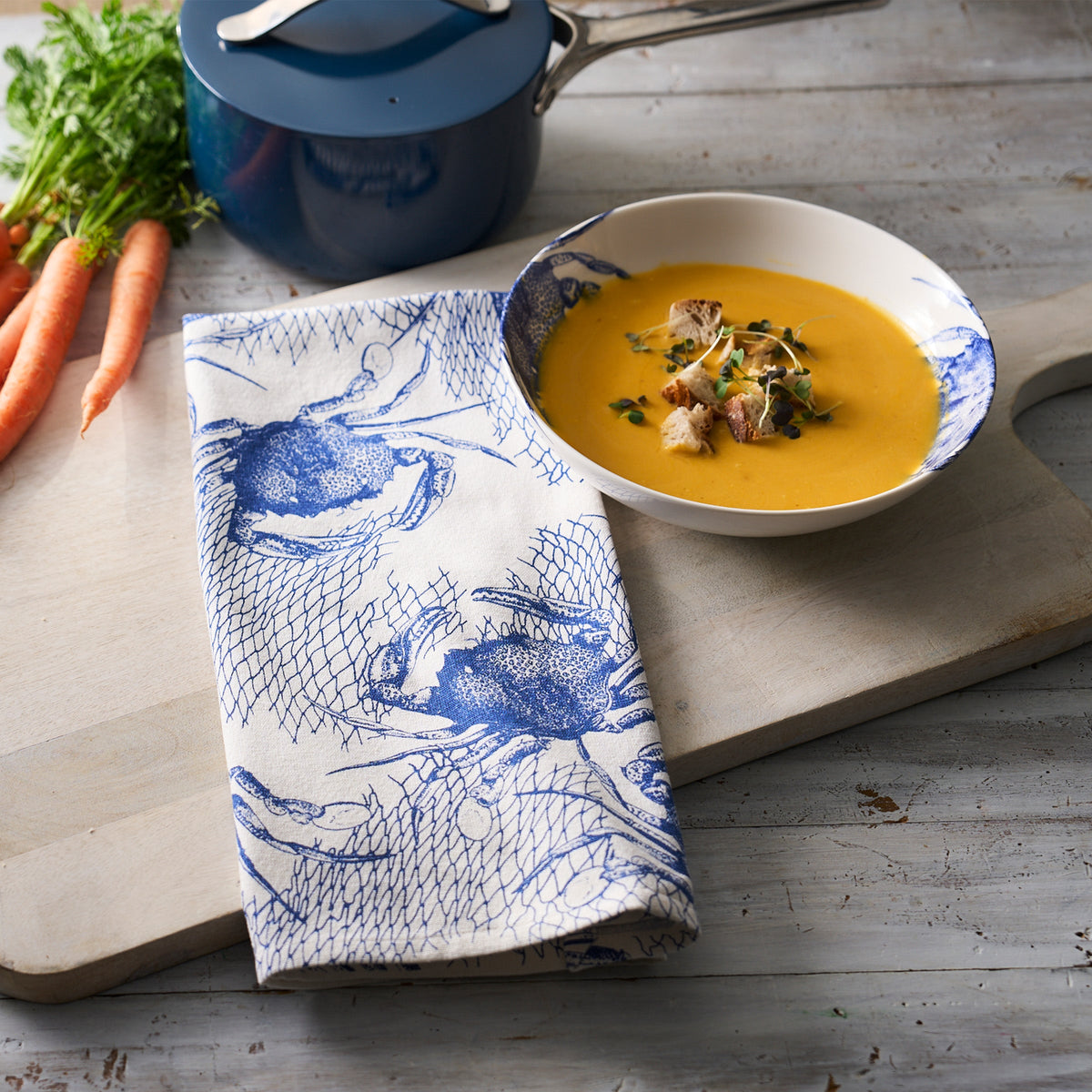A bowl of soup with carrots on a cutting board, adding a touch of seafaring whimsy, draped with the Crabs &amp; Nets Kitchen Towels Set/2 by Caskata.