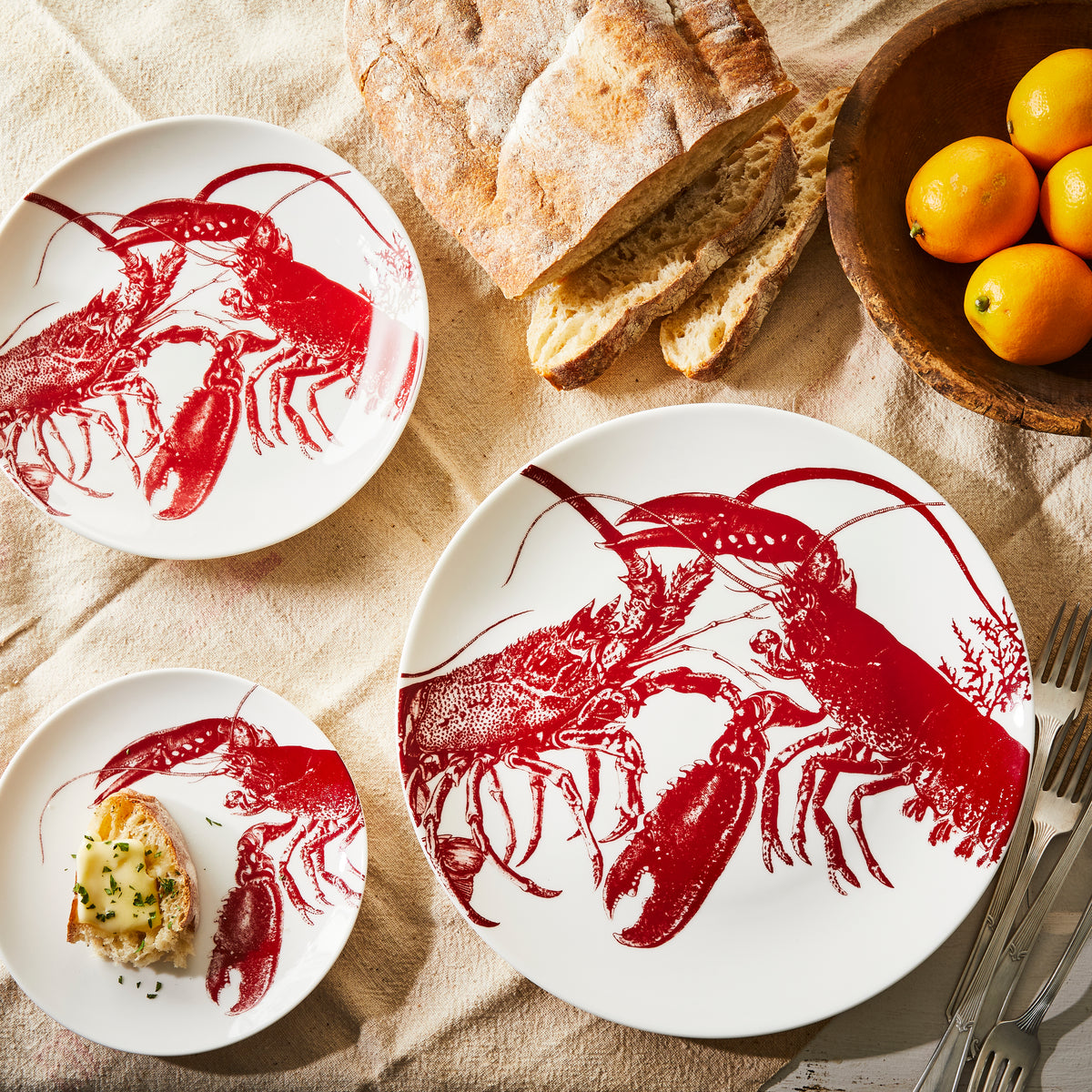 A set of three Caskata Artisanal Home Lobster Red Small Plates with red designs, a bowl of oranges, bread, and a small dish with bread and spread are arranged on a cream tablecloth with silverware nearby, evoking a seaside style.