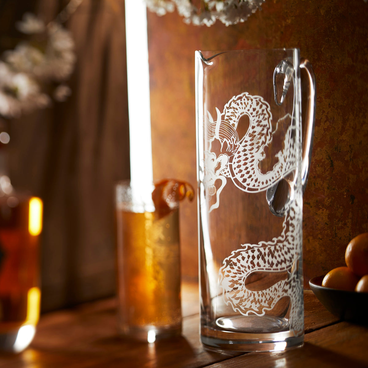The sand etched dragon pitcher by Caskata is displayed on a moody warm tabletop with  coordinating tall drinking glass.