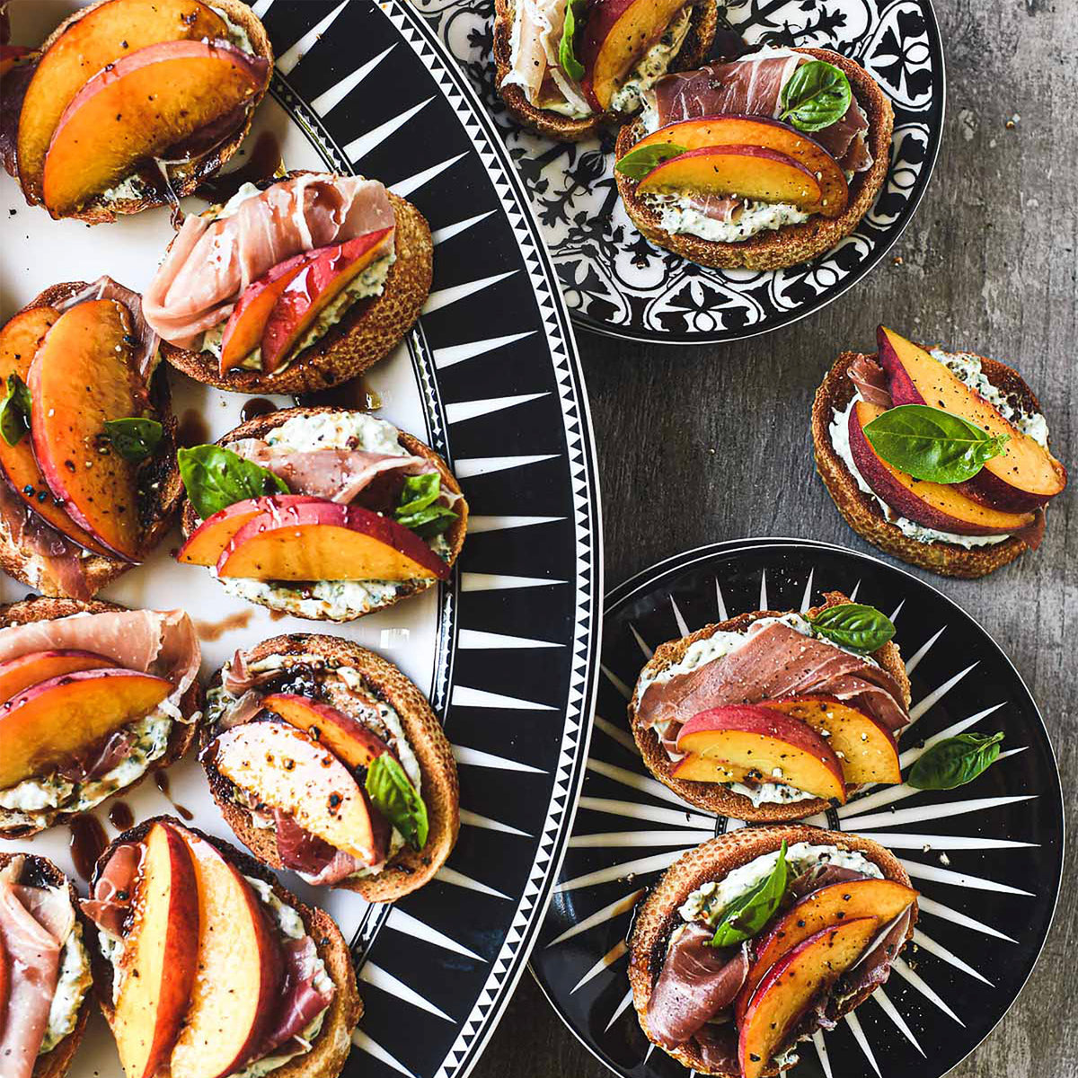 A variety of appetizers featuring sliced peaches, prosciutto, ricotta, and basil on toasted bread are elegantly presented on the Marrakech Oval Rimmed Platter from Caskata Artisanal Home.
