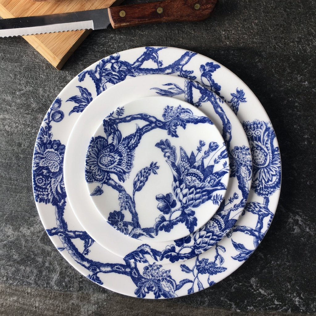 Three white ceramic plates with intricate blue graphic florals, stacked on a gray surface next to a wooden cutting board and a serrated knife with a wooden handle. This premium Arcadia Rimmed Dinner Plate by Caskata Artisanal Home feels like it should be displayed in the collections of the Williamsburg Foundation.