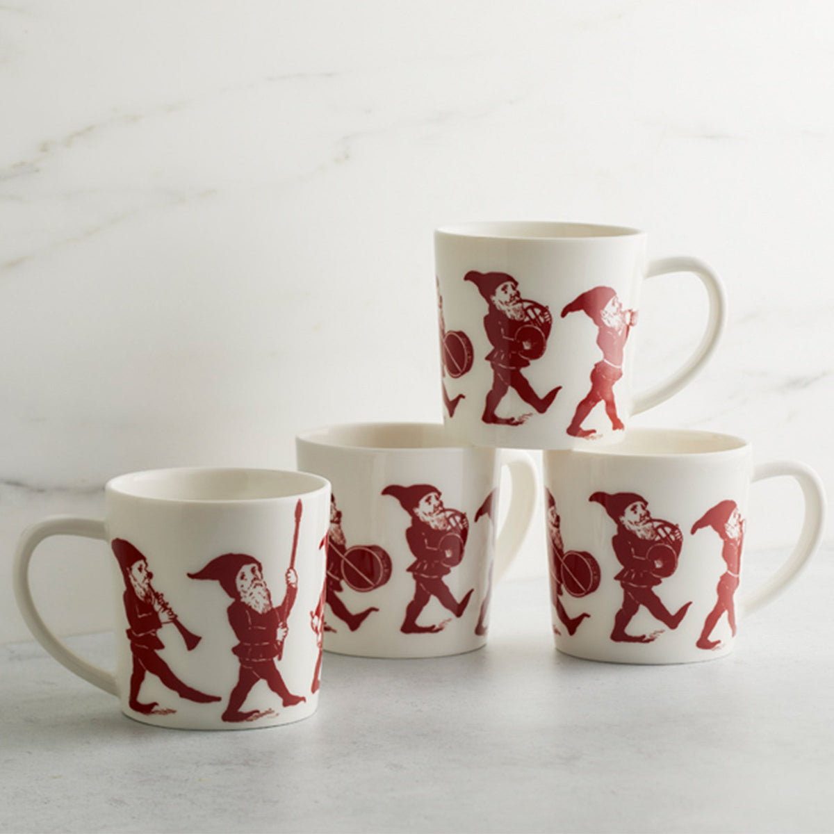 Four porcelain &quot;Elves Mug&quot; by Caskata Artisanal Home with red illustrations of playful elves, stacked in a pyramid formation, placed on a white surface with a marble background. This delightful holiday collection adds a whimsical touch to any festive setting.