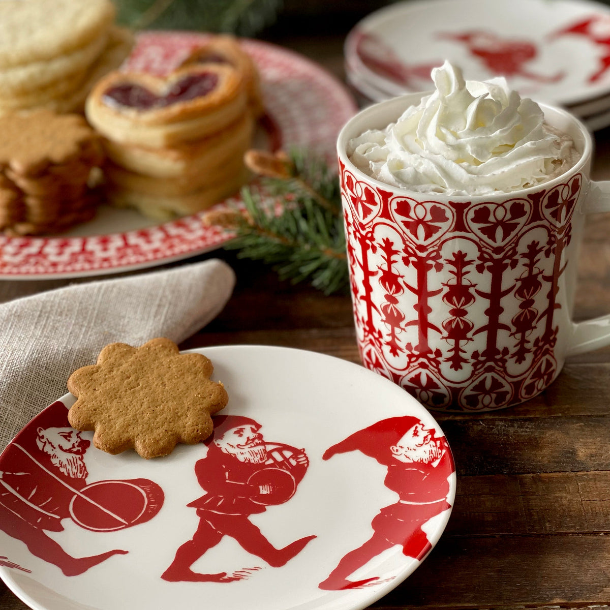 A holiday-themed table setting with a gingerbread cookie on a plate, a mug of whipped cream-topped hot beverage, and a background of assorted cookies. Elves Small Plates by Caskata Artisanal Home from our holiday collection feature festive red designs, making your table an entertaining powerhouse perfect for any gathering.