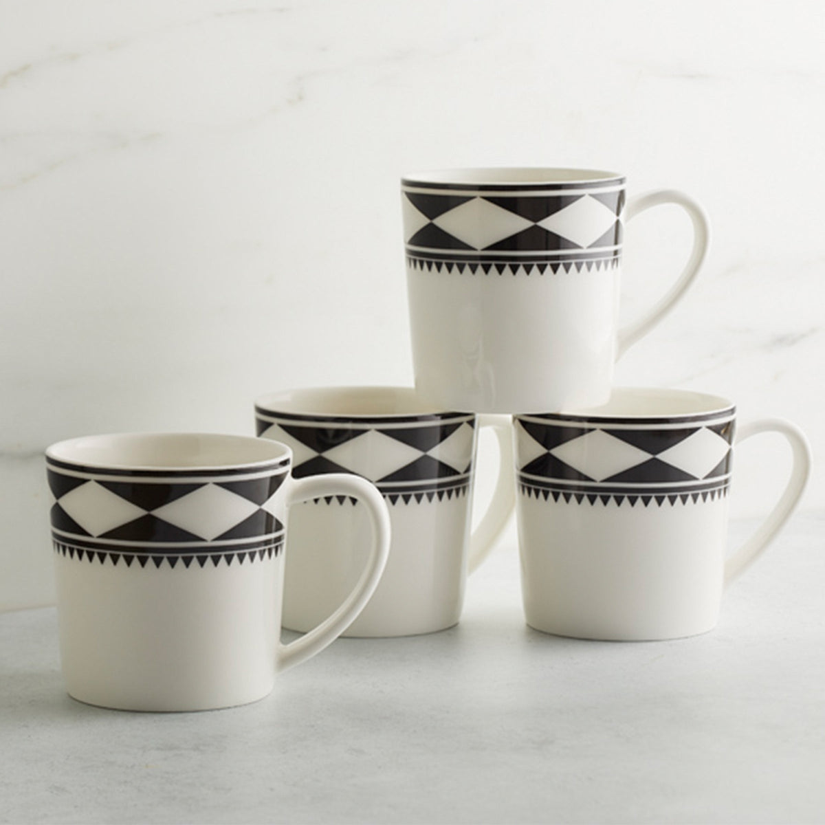 Four white Fez Mugs with black geometric patterns, inspired by Moroccan designs, are stacked on a gray surface against a white marble background. Made from high-fired porcelain by Caskata Artisanal Home, these beautiful mugs are both dishwasher and microwave safe.