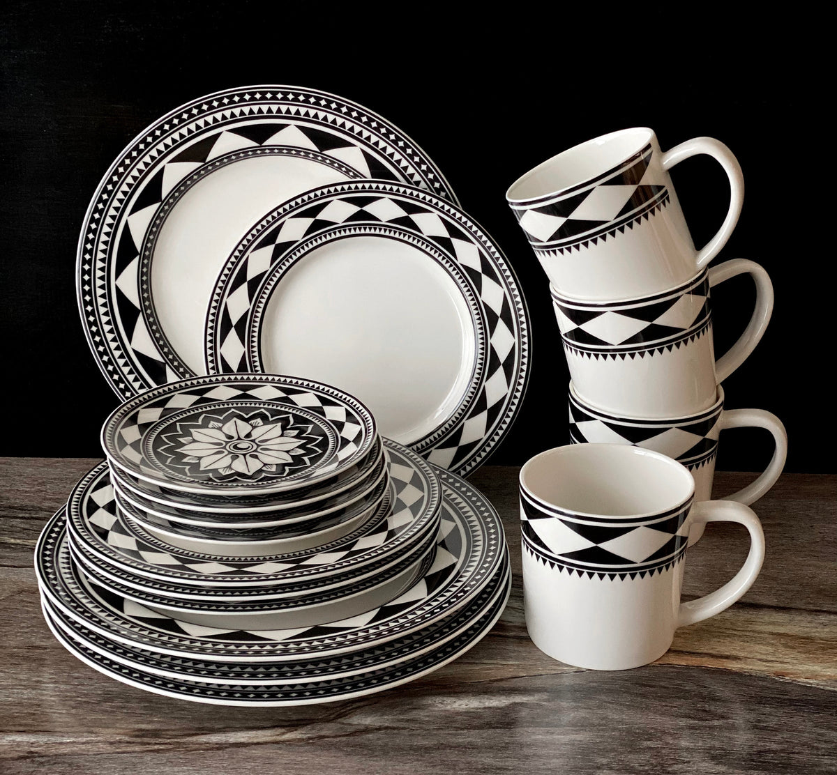 A set of black and white geometric patterned dinnerware including plates, bowls, and a Fez Mug from Caskata Artisanal Home, all made from high-fired porcelain and neatly stacked on a wooden surface. Dishwasher and microwave safe.
