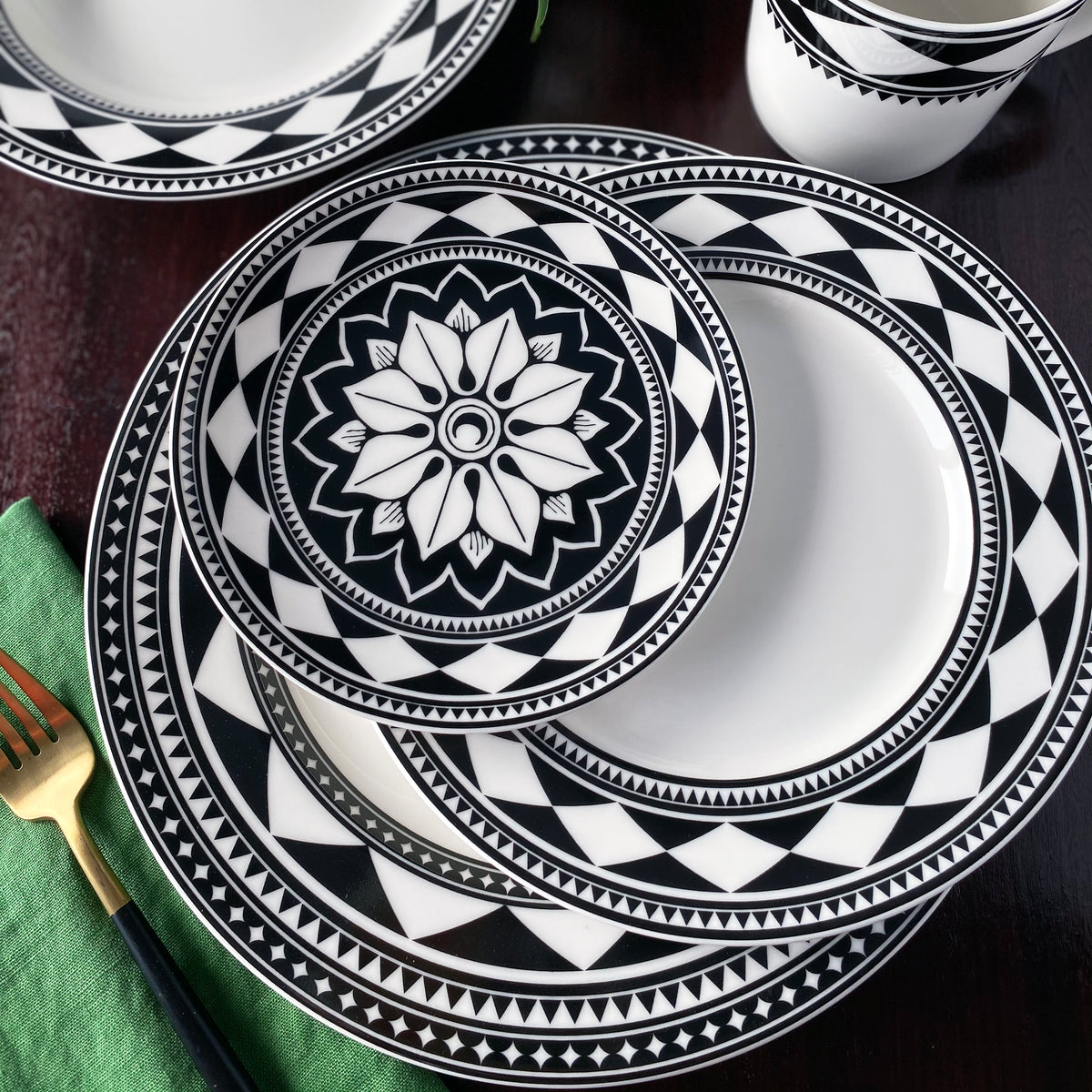 A set of sophisticated dinnerware with Caskata Artisanal Home&#39;s Fez Rimmed Dinner Plate, featuring black and white geometric patterned plates and bowls arranged on a dark brown table, accompanied by a green napkin and a fork.