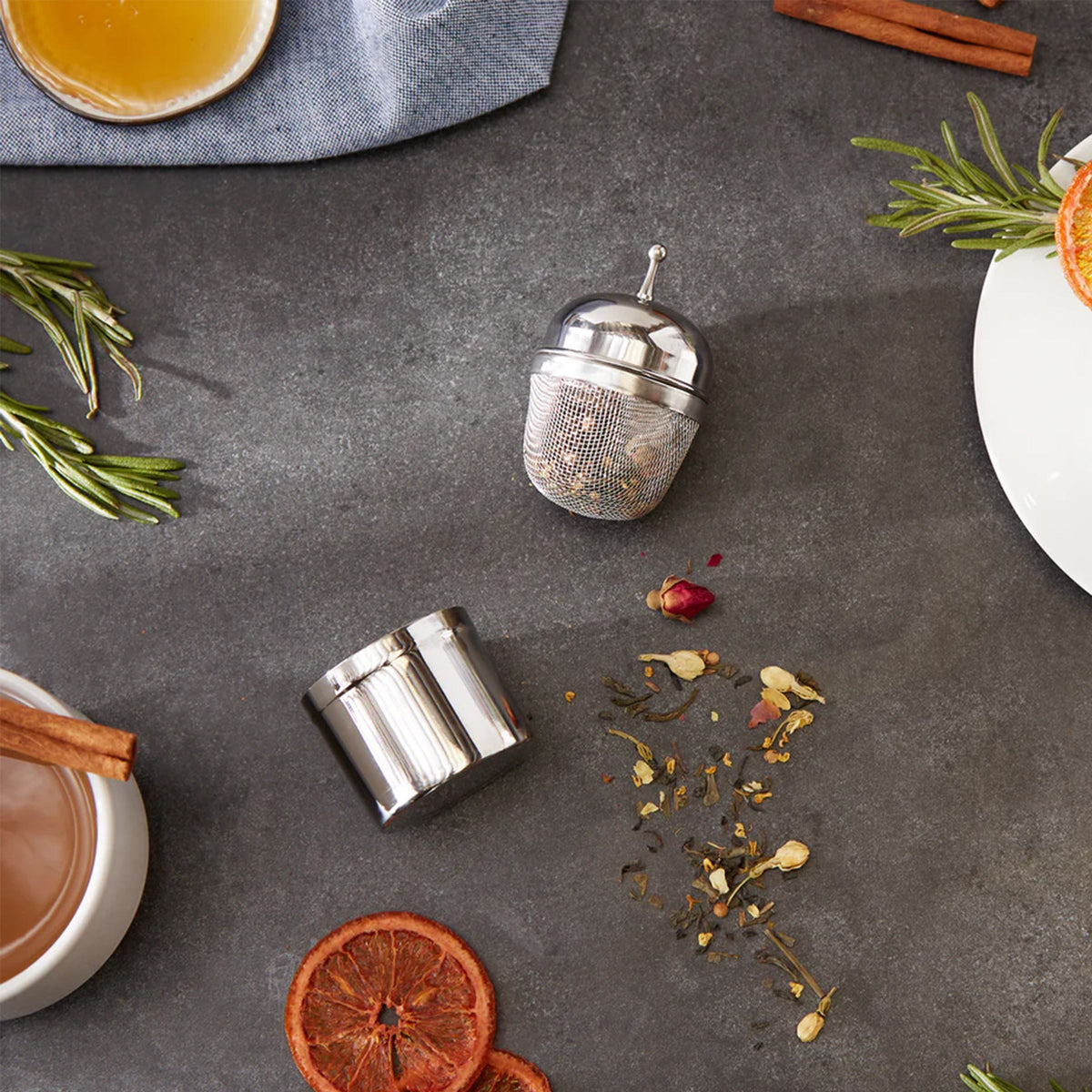 A RSVP International stainless steel reusable Floating Tea Infuser &amp; Caddy steeping loose leaf tea, complemented by cinnamon sticks and oranges on a table.