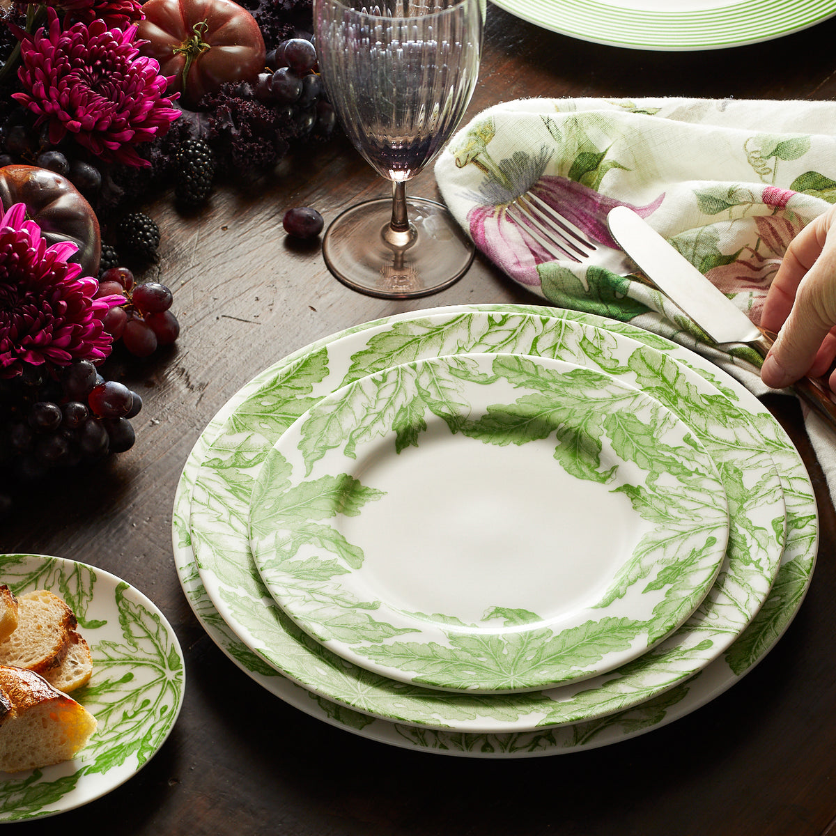 A table setting features green and white leaf-patterned plates, a wine glass, bread, and a floral napkin. A hand places a knife beside the Caskata Artisanal Home Freya Rimmed Charger Plate. Purple flowers and grapes are in the background, evoking an atmosphere worthy of the goddess of love.